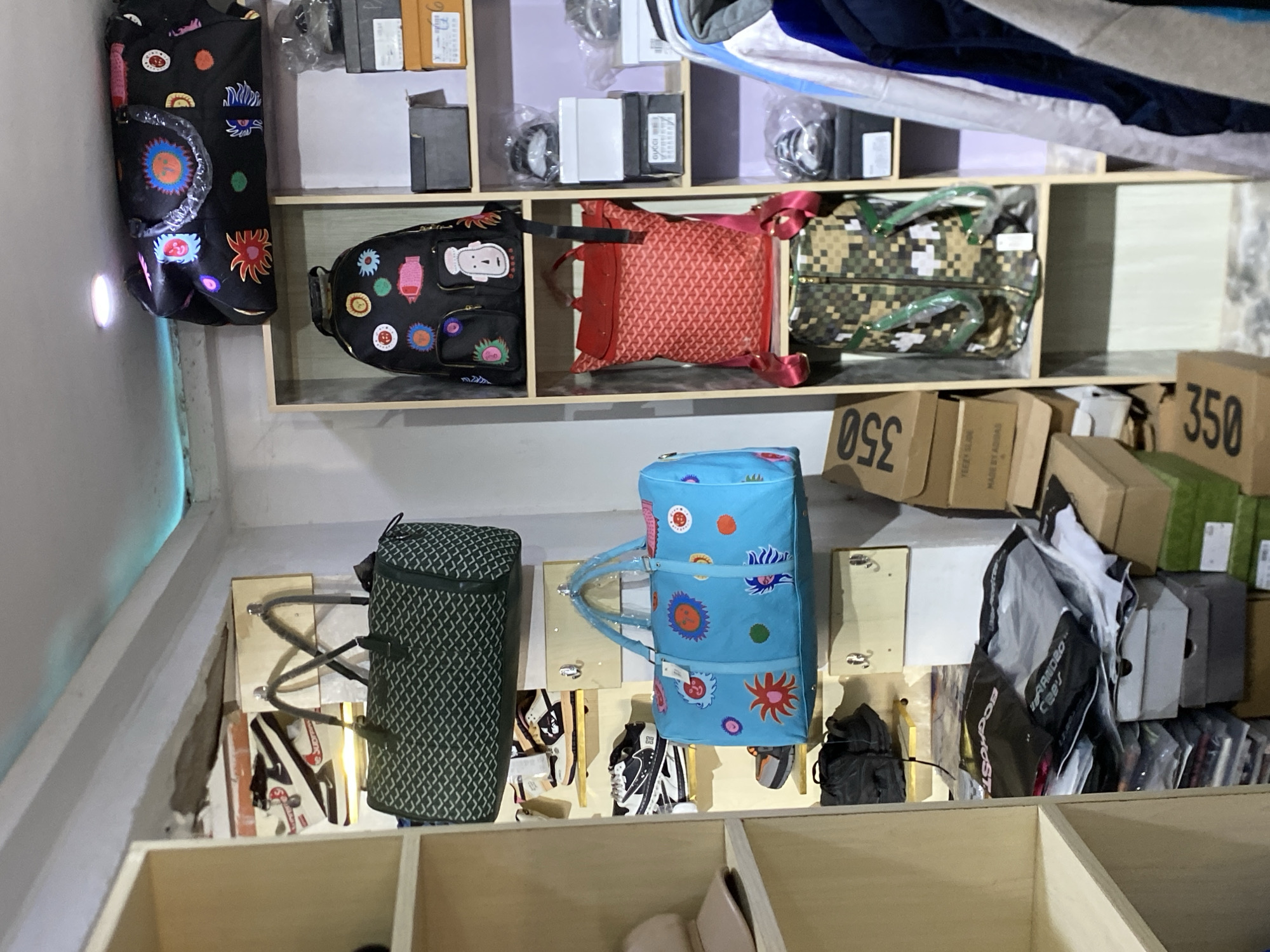 The image shows a storage area, likely a retail or storage space, in an indeterminate location, but supposedly in Bukuru, Nigeria. There are several shelves holding bags, shoeboxes, and other merchandise.
Top shelf: A black bag with colorful designs hangs on the left. Beside it, a black bag with cartoonish faces, a red patterned bag, and a camouflage pattern bag are on display. Small boxes and packing materials are also visible on the top shelf.
Middle shelves: Two cylindrical bags, one green and one blue with colorful designs, stand on the lower shelf. Shoes are hanging on hooks or pegs below them. To the right, there are shoeboxes stacked, some with the word "Adidas" visible. Cardboard boxes labelled "350" are present on top of the shoeboxes.
Bottom shelves: Below the middle shelves, there are empty rectangular compartments.