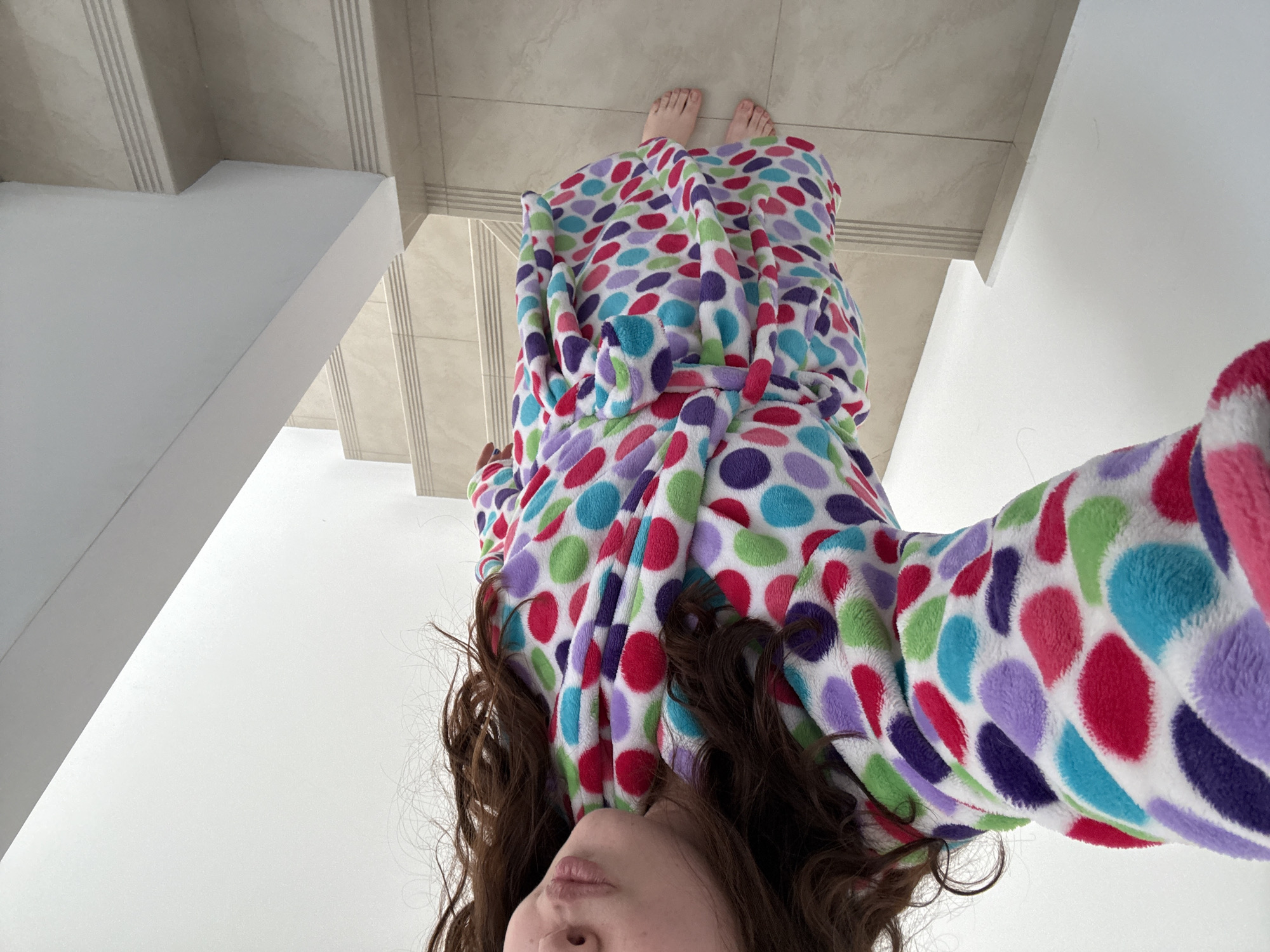 A woman in a colorful polka-dot bathrobe is upside down, her feet visible near a staircase and tile floor. Her brown hair hangs down, and part of her face is visible. The location appears to be indoors, possibly a residence. The city of Santa Clara Ocoyucan, Mexico, is not discernible from the image alone.