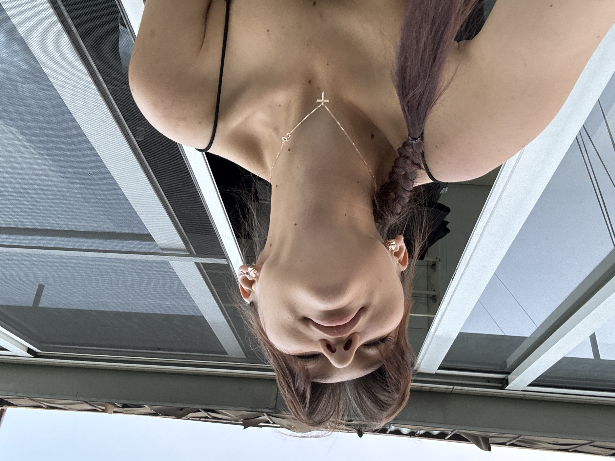 The image shows a young woman with her head tilted to the side, looking up. She has long, brown hair that is braided and falls over her shoulder. She is wearing a thin strap top and a delicate necklace with a cross pendant. Her ears are adorned with small, gold earrings. The background appears to be a window with a screen, suggesting an indoor or balcony setting. The lighting is soft and natural. The overall tone of the image is casual and intimate. There is no discernible information about the location being Novokuznetsk, Russia.