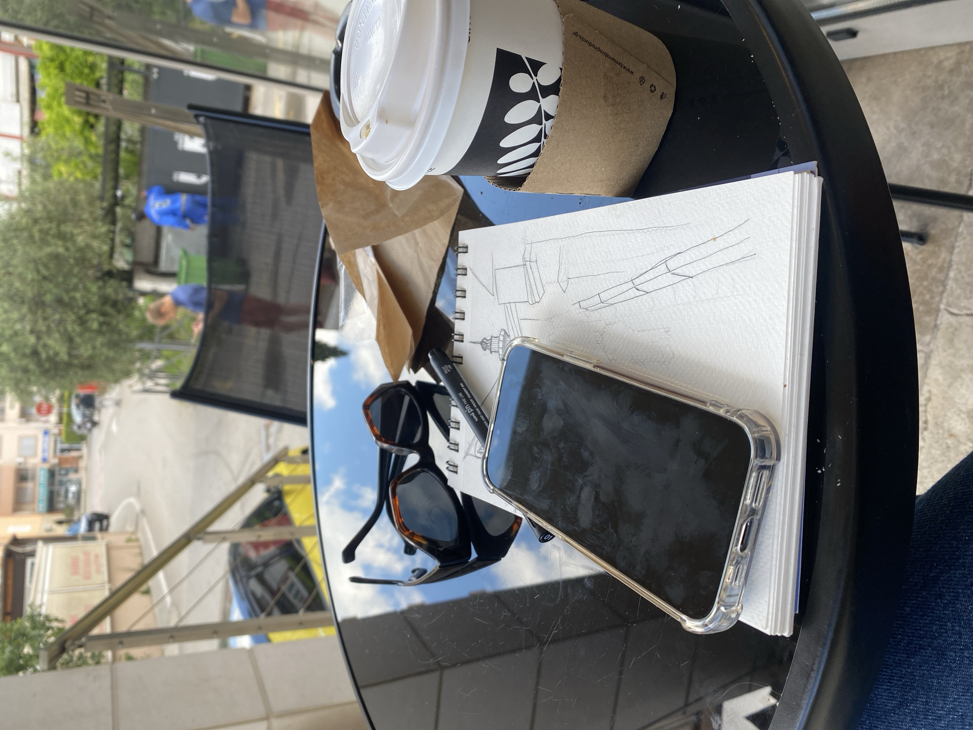 A black table sits outside, holding a cup of coffee in a cardboard sleeve, a sketchbook with a drawing on it, a black marker, a mobile phone, and a pair of sunglasses. Reflections of the sky are visible on the glossy table surface. In the background, a street scene is visible with buildings, parked cars, trees, and people.