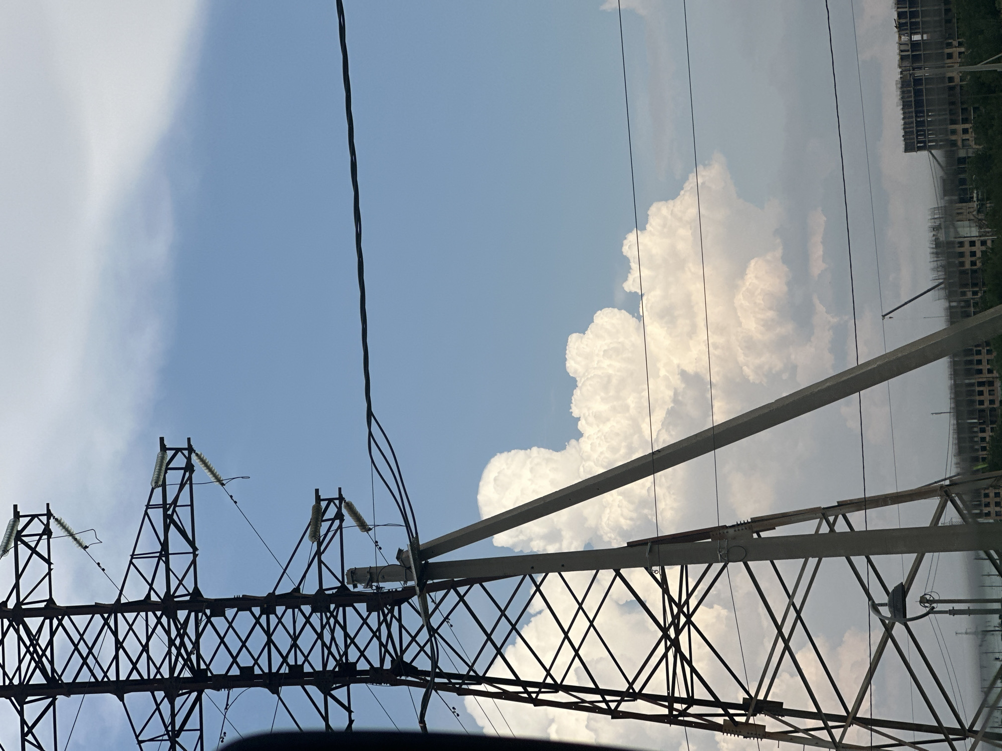 The image captures a view of a high-voltage power line tower against a partly cloudy sky. The tower is constructed of dark metal lattice and is accompanied by a concrete support structure. Several power lines stretch horizontally across the frame. In the background, there are construction sites with buildings under development and some trees. The scene appears to be an outdoor, urban or semi-urban environment, likely in the city of Krasnodar, Russia, given the context. There are no people or animals visible in the image. The lighting suggests it is daytime.