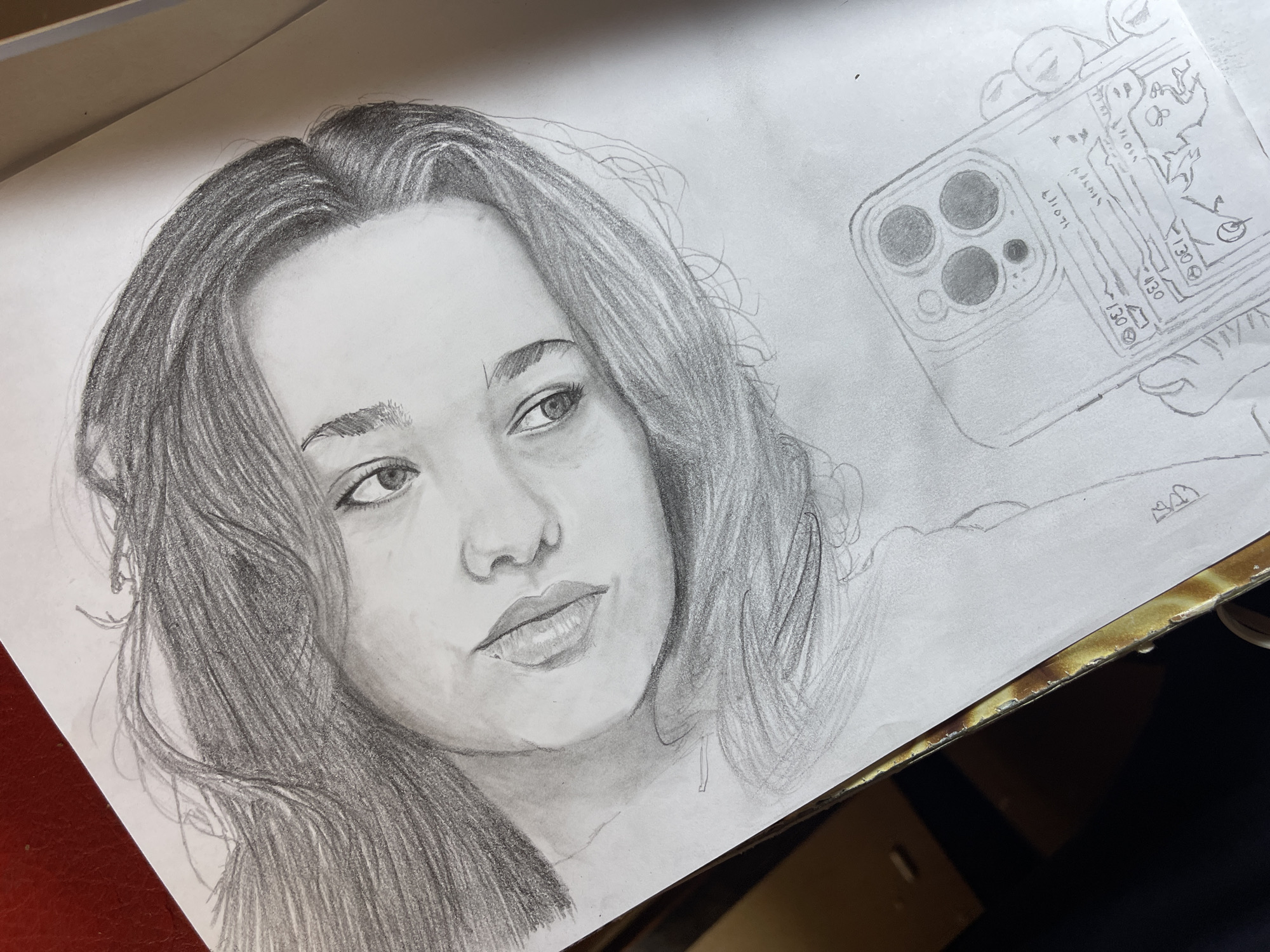 The image is a pencil drawing on white paper. The drawing depicts a portrait of a young woman holding a cell phone up in a selfie pose. The woman has medium-length wavy hair, and is looking off to the side with a slightly pensive expression. The phone appears to be an iPhone with three camera lenses. The background is plain. The drawing appears to be laid on a table near a red surface and some dark material that could be a desktop or other surface. There are no visible indications that the image originates from Jos, Nigeria.