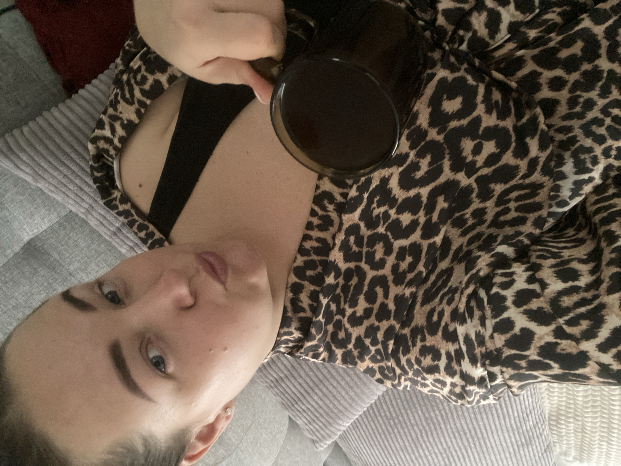 The image shows a woman lying on a gray corduroy-textured surface, possibly a couch or bed. She is wearing a leopard-print top over what appears to be a black bra or tank top. She holds a black mug filled with a dark liquid, likely coffee. The woman has fair skin, dark eyebrows, and blue eyes. Her hair is pulled back. The background is blurred, but there are hints of a reddish-brown surface behind the gray corduroy. The image gives no indication of a location in Sochi, Russia or any other specific place. The image captures a personal, casual moment.