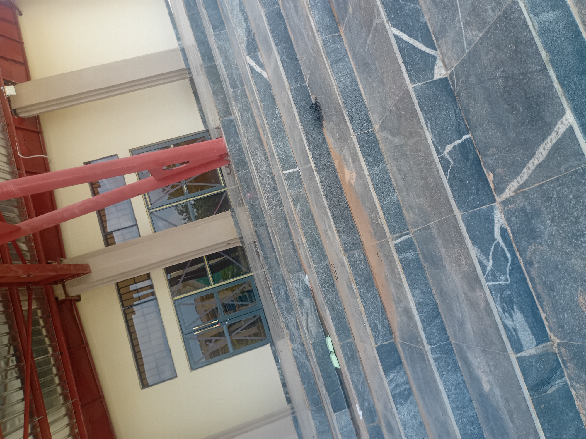 The image shows a wide outdoor staircase made of dark grey stone tiles with lighter veining. The stairs are illuminated by natural daylight. The staircase leads up to a building with a beige facade. Above the entrance, there are large glass windows with dark frames, and to the left of the entrance, there are two smaller, horizontal windows. A red steel beam is prominently visible supporting the roof structure, which appears to be made of corrugated metal. The ground at the base of the stairs is covered with loose dirt and small debris. There are no people visible in the scene. The image is taken from a low angle, looking up the stairs.