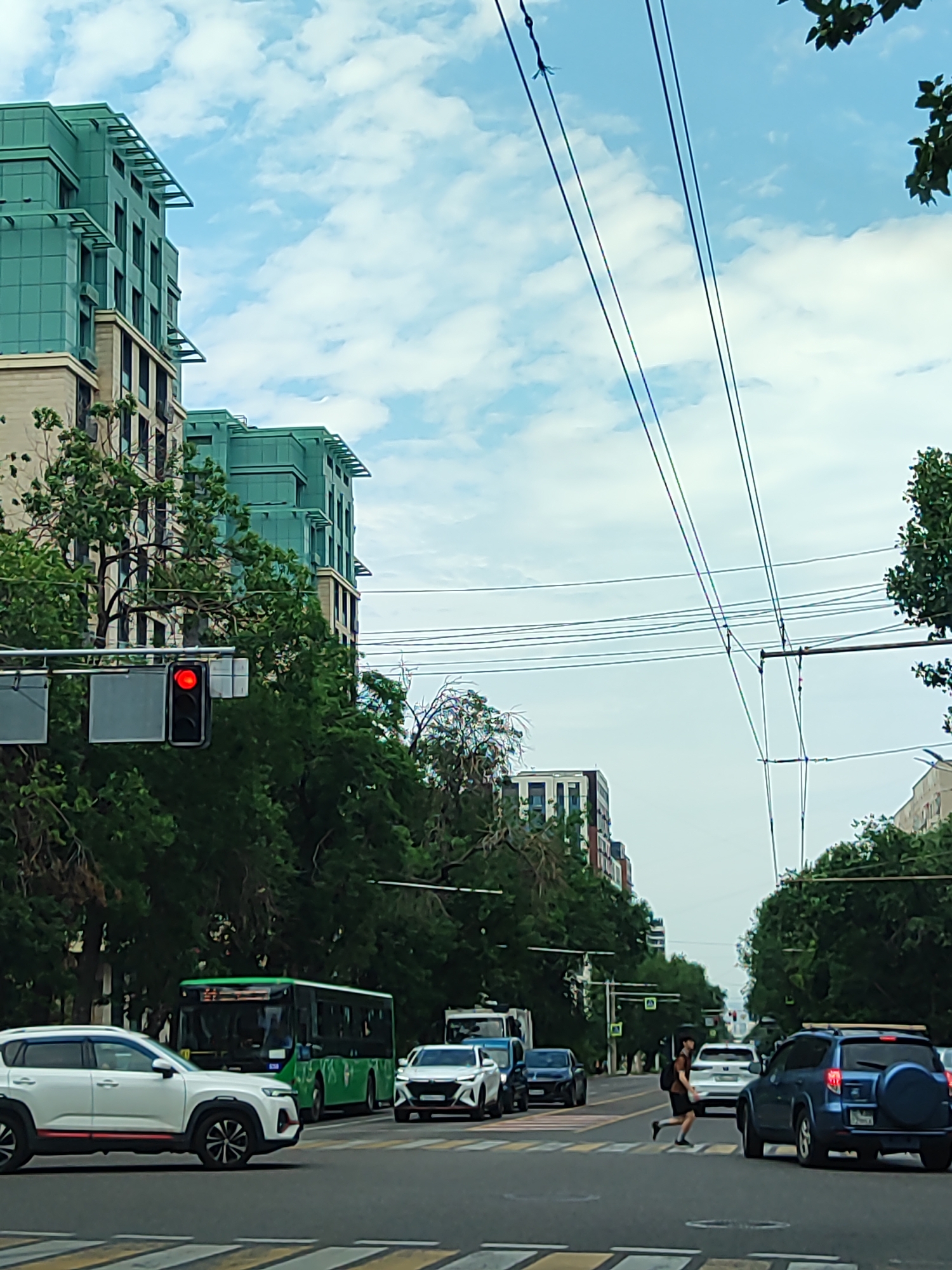 The image captures a street scene in Almaty, Kazakhstan. A prominent modern building with teal-colored facade elements stands on the left, dwarfing the lush green trees lining the street. A traffic light with a red signal is visible on the left. Several vehicles, including a white SUV, a green bus, and a blue SUV, are present on the road. A pedestrian is crossing the street in the middle distance. Overhead power lines crisscross the sky, indicating an urban environment with public transportation or utility infrastructure. The sky is partially cloudy, suggesting daytime. The overall impression is a busy city intersection with a mix of architecture and daily life.