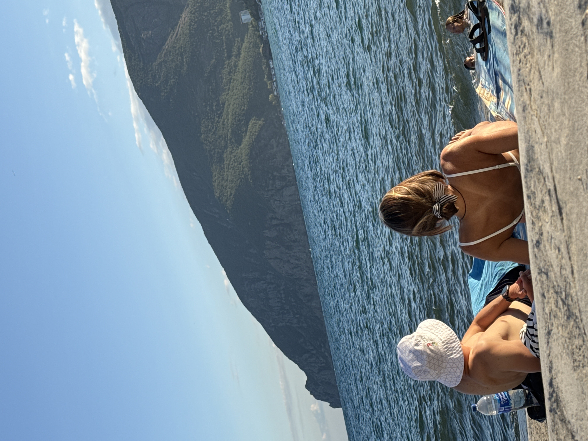 In a serene coastal scene, two individuals are seated on a beach with their backs to the camera, facing the shimmering blue ocean and a verdant, mountainous landscape in the distance. The man, on the left, wears a white bucket hat and rests his arm on a blue towel. A plastic water bottle sits beside him. The woman, on the right, is in a white bikini, her hair tied back with a striped ribbon. In the background, several people are visible in the water. The sky above is a clear, bright blue with a few wispy clouds. The environment suggests a tranquil day at the beach, possibly in the Crimean Peninsula of Ukraine, given the distinctive mountain formation and coastal architecture.