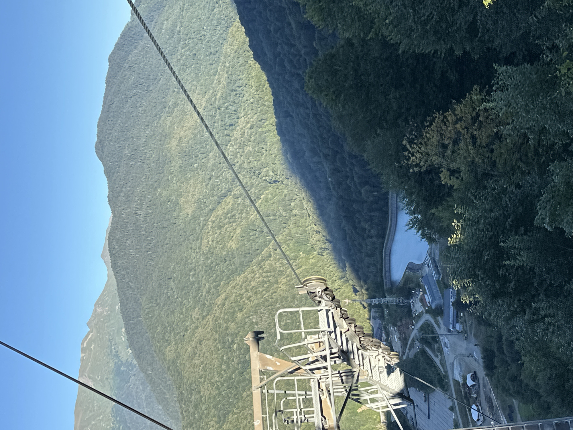 The image, taken from a ski lift, captures a panoramic view of the mountain landscape in Estosadok, Russia. The foreground features the intricate machinery of the ski lift, including cables, pulleys, and support structures, all coated in a weathered, metallic finish. Beyond the lift, the scene unfolds into a series of densely forested mountains. Sunlight casts a dramatic shadow across one of the slopes, creating a stark contrast between illuminated and shaded areas. In the middle ground, a clear blue reservoir is visible, bordered by a few small buildings and industrial structures, suggesting infrastructure related to the ski resort or water management. The background is dominated by more distant, softer mountain peaks under a clear blue sky. The overall impression is one of natural beauty integrated with man-made structures, characteristic of a mountain resort area.