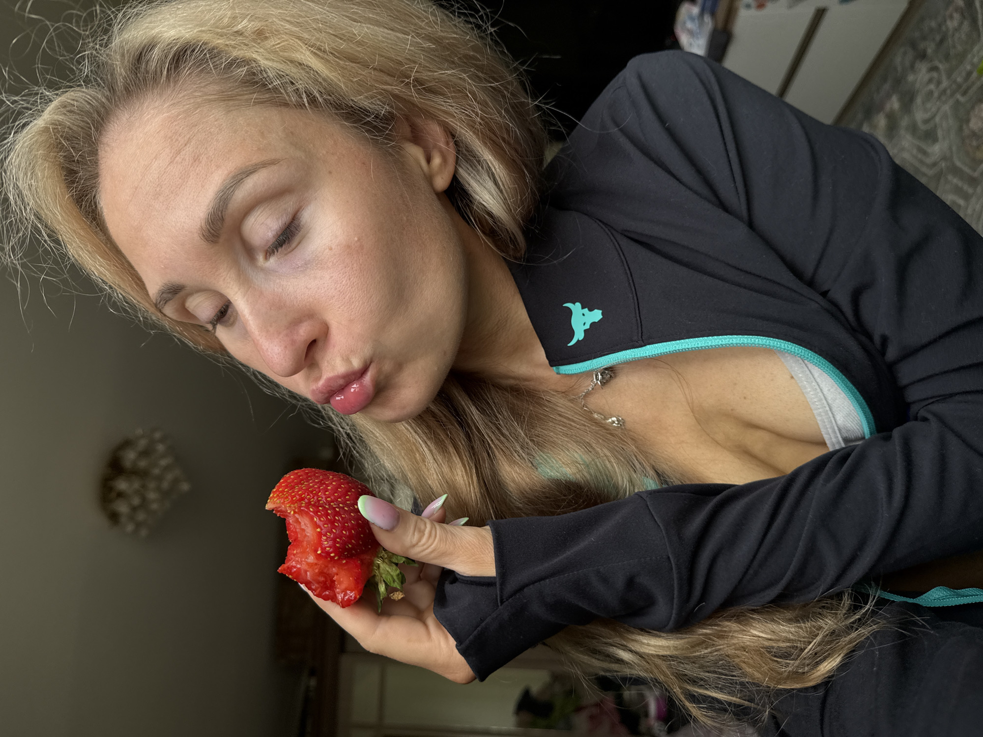 The image shows a close-up of a blonde woman holding a strawberry near her lips, which are pursed as if about to kiss the fruit. She is wearing a black zip-up jacket with teal trim and a teal bull logo on the left chest. The background is dimly lit and appears to be an indoor setting, possibly a home. There is a chandelier visible in the upper portion of the image. The woman has blonde hair and her eyes are closed. The strawberry is partially eaten. No specific location in Chaikovsky, Russia is identifiable from the visual information in the image.