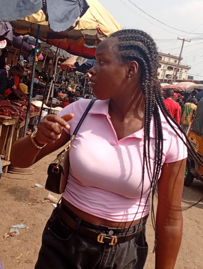 The image captures an adult female, likely of African descent, standing in an outdoor market in Onitsha, Nigeria. She is positioned slightly right of center, looking towards the right, with dark cornrow braids extending into long, loose individual braids. She wears a light pink, short-sleeved polo shirt, dark jeans, and a dark belt with a gold-colored, stylized buckle. A dark shoulder bag with a gold chain strap is carried over her left shoulder, and a gold-colored bracelet adorns her left wrist. Her left hand is partially raised, fingers slightly curled.
The background reveals a densely packed market scene with multiple stalls shaded by large, colorful umbrellas (blue, yellow, orange, red). To the left, a stall displays cuts of raw meat on a wooden table, with a male vendor partially visible behind it. Various goods, including what appear to be clothing and packaged items, are visible on other stalls further back. In the distant background on the right, a multi-story building with balconies is discernible under a bright sky, alongside utility poles and wires. The ground is unpaved dirt, showing scattered debris.