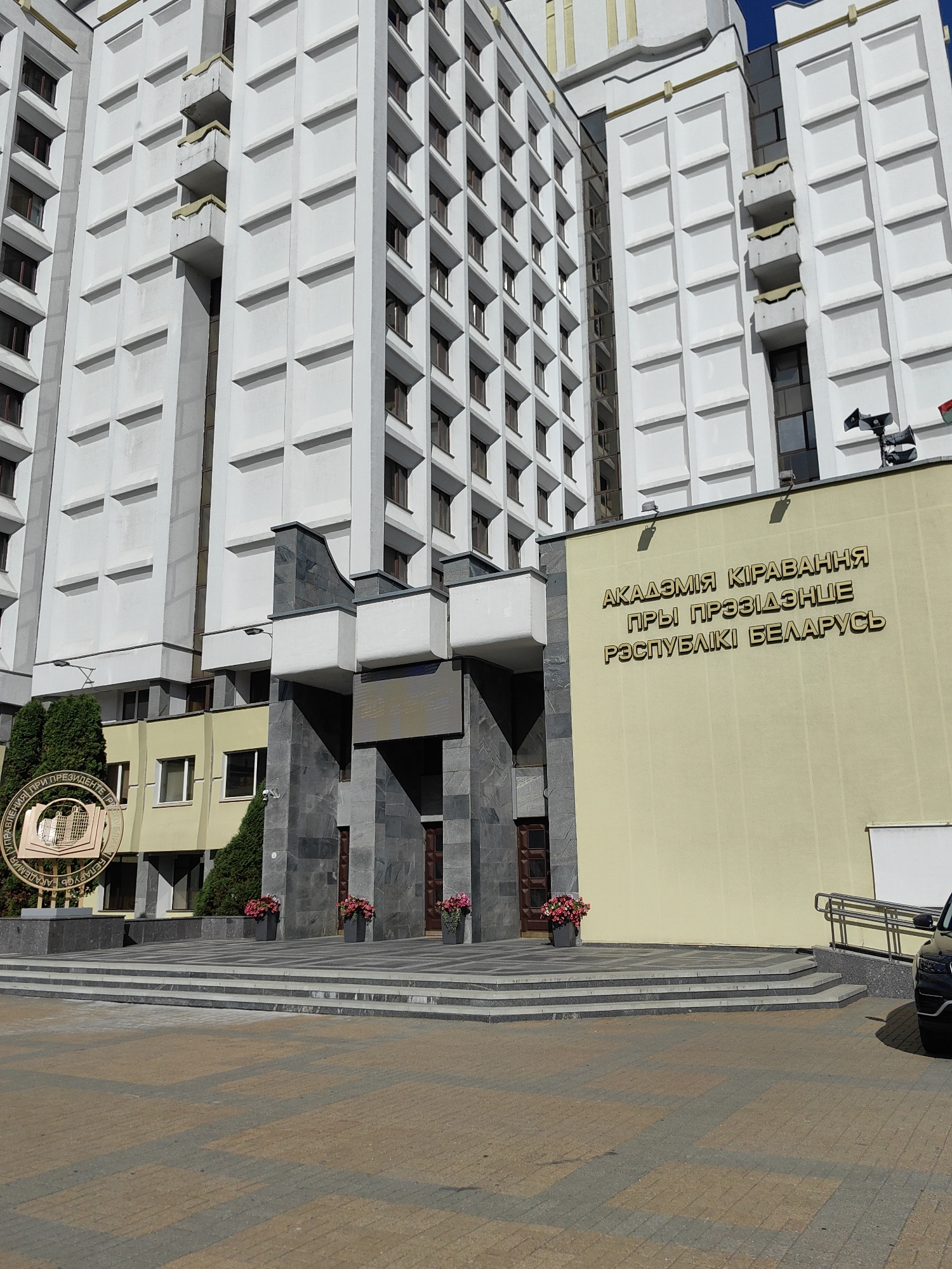The image shows the Academy of Management under the President of the Republic of Belarus, located in Minsk, Belarus. The building is a large, modern structure with a white facade and multiple windows. The entrance to the building is prominent, with a set of wide stairs leading up to it. Above the entrance, there is text in Cyrillic which translates to "Academy of Management under the President of the Republic of Belarus." There are also decorative elements such as planters with flowers and some signage. A car is partially visible on the right side of the image. The ground in front of the building is paved with a patterned stone. The sky is clear and blue.