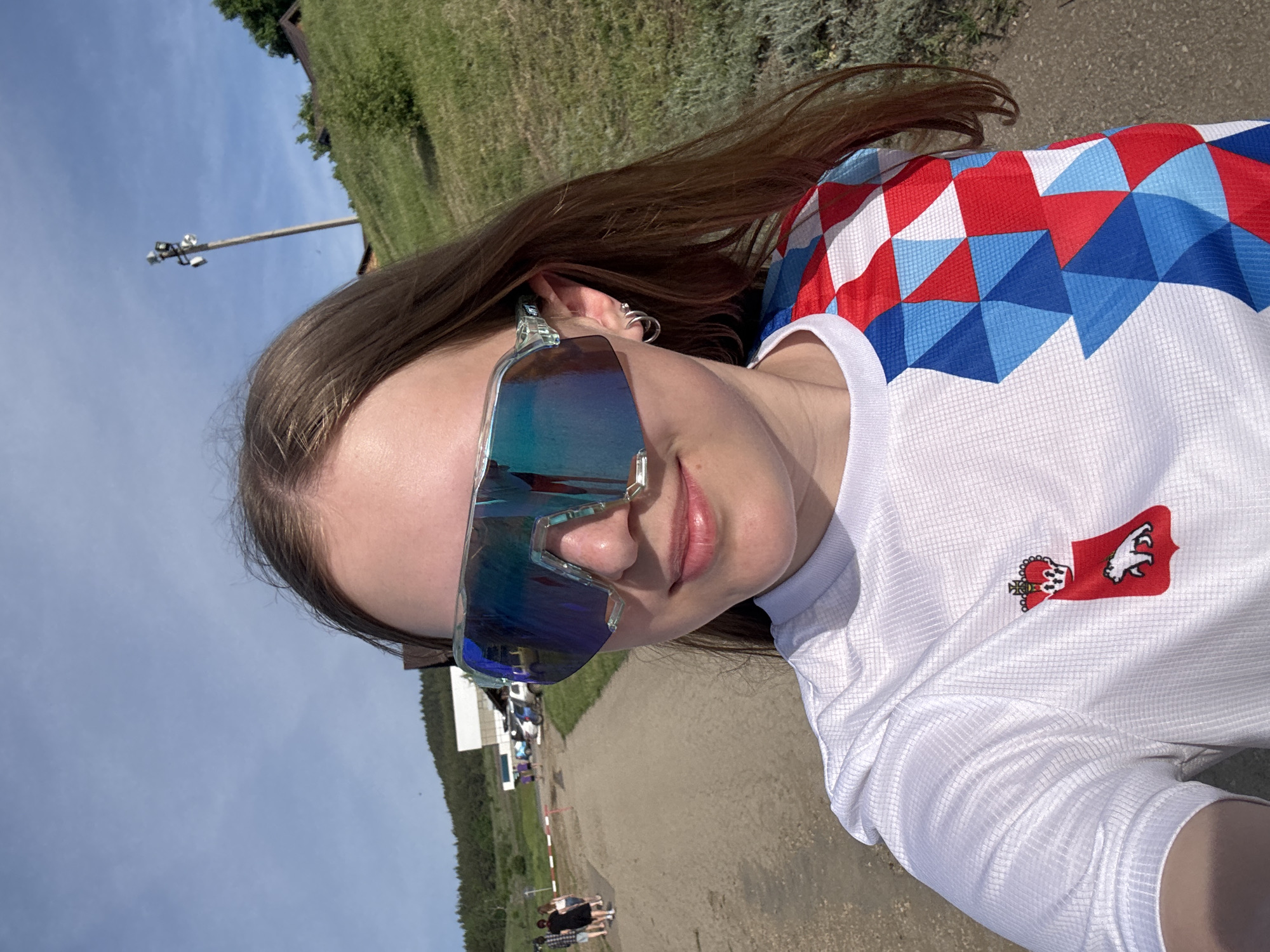 A young woman is pictured outdoors, wearing blue mirrored sports sunglasses and a white shirt with a pattern of red, white, and blue geometric shapes across the shoulders. The shirt also has a red patch on the lower right featuring a white bear with a crown. The woman has long brown hair.
The background appears to be a park area. Behind her, a pathway leads towards buildings in the distance. There are trees and grassy slopes on either side of the path, and what appears to be a lamppost on the left. In the distance, there are a few people walking. Based on the patch on her shirt, the picture was likely taken in the city of Khvalynsk, Russia.