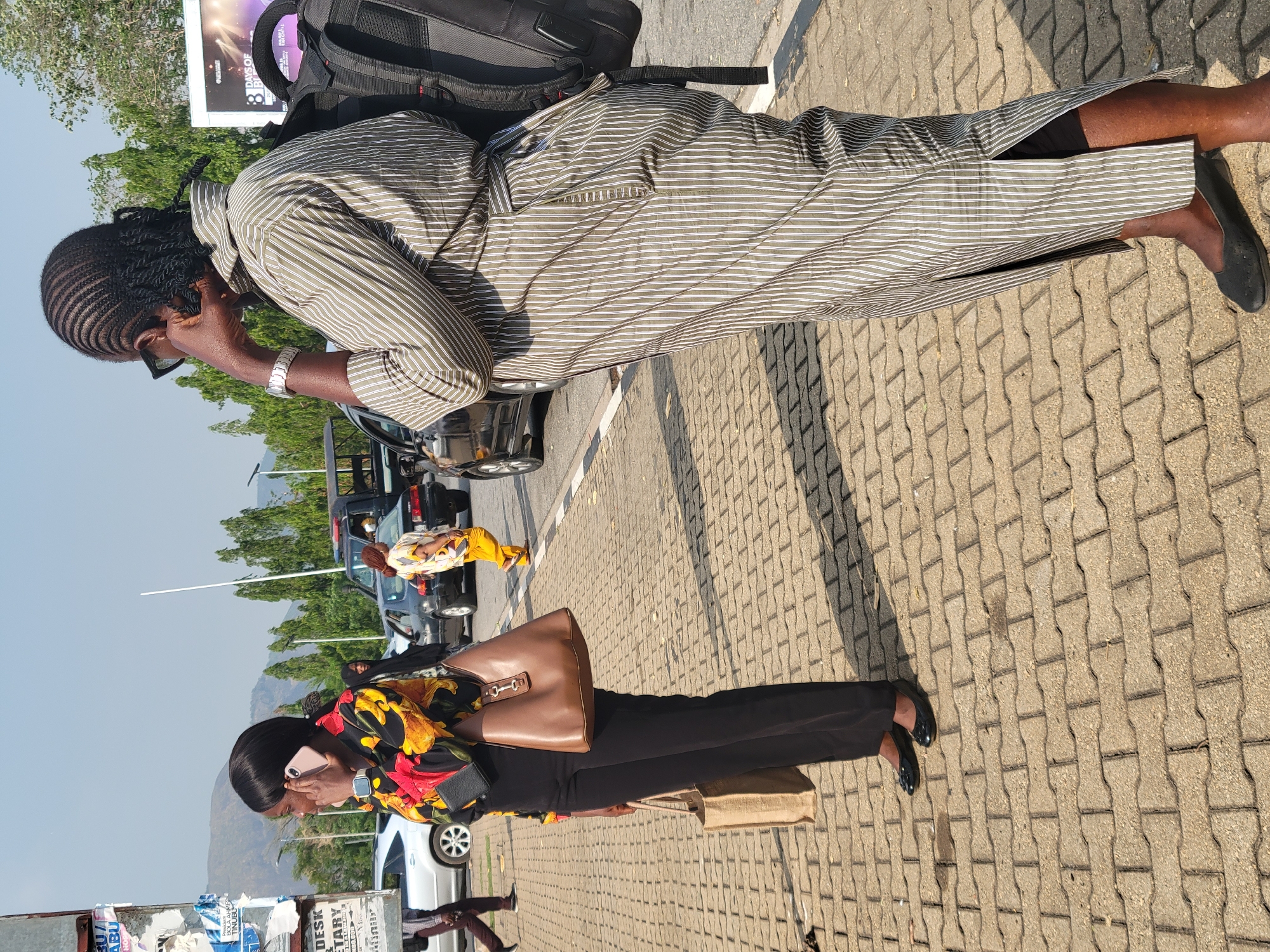 Two women are standing on a paved sidewalk in Abuja, Nigeria.
The first woman, closer to the camera, is wearing a colorful floral top, black pants, and black shoes. She is holding a large brown purse and a smaller tan bag. She is holding a pink cell phone to her ear.
The second woman, further away, is wearing a striped dress and black shoes. She has a black backpack on her back. She is holding a cell phone to her ear.
Several cars are parked on the street behind the women. A third person in yellow and white clothing is beside one of the cars. There are trees and a hill in the background. There are utility poles and a billboard in the background. The sky is clear and blue.