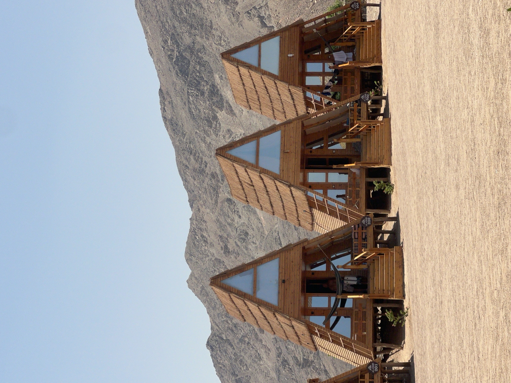This is a daylight, eye-level, outdoor, wide shot of a row of triangular, wooden cabins in a desert landscape near Nueiba, Egypt. The cabins have thatched roofs and glass triangular windows at the top. Each cabin is elevated on stilts and has wooden stairs leading up to a front porch. Some cabins have hammocks or clothes hanging from their porches. The ground is sandy, and a rocky, barren mountain range is visible in the background under a clear blue sky. The cabins are numbered, with visible numbers including 201, 202, and 204, suggesting a resort or lodging facility. One person is partially visible standing in the doorway of the second cabin from the left.
