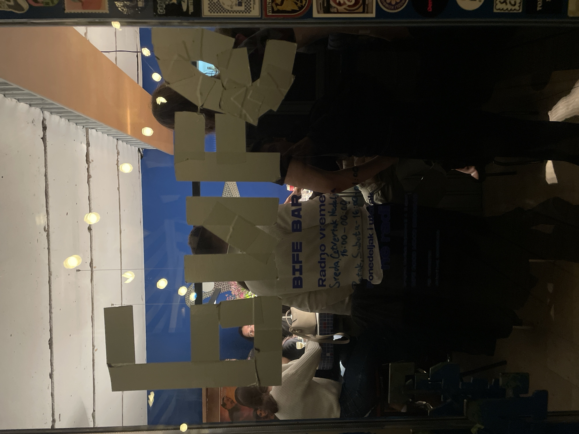 The image depicts the interior of Bife Bar, located at Karađorđeva 57, 11000 Belgrade, Serbia, viewed through its glass entrance. Large, light-colored block letters spelling "BIFE BAR" are affixed to the glass, partially obscuring the interior view. Below this, operating hours are displayed in Serbian script: "Radno vreme: Sreda, Četvrtak, Nedelja 16:00-00:00, Petak, Subota 16:00-01:00, Ponedeljak i utorak ne radi" (Working hours: Wednesday, Thursday, Sunday 16:00-00:00, Friday, Saturday 16:00-01:00, Monday and Tuesday closed). A vertical blue frame on the far right is adorned with multiple stickers.
Inside the establishment, approximately five individuals are seated at tables. The lighting is provided by several warm, circular pendant lights hanging from a light-colored ceiling. The background wall is painted a distinct deep blue. One individual in a light-colored long-sleeved top is seated at a table on the left, while another in a similar top is seated with their back to the camera, partially blocking the bar's name. A person with dark hair and a dark top is visible looking towards the right. On a table, a light-colored handbag and a glass containing a pale liquid are visible. An artistic mosaic-like piece is present on the blue wall, along with a framed image to its left. People are observed in a social setting, appearing to be engaged in conversation or relaxation.