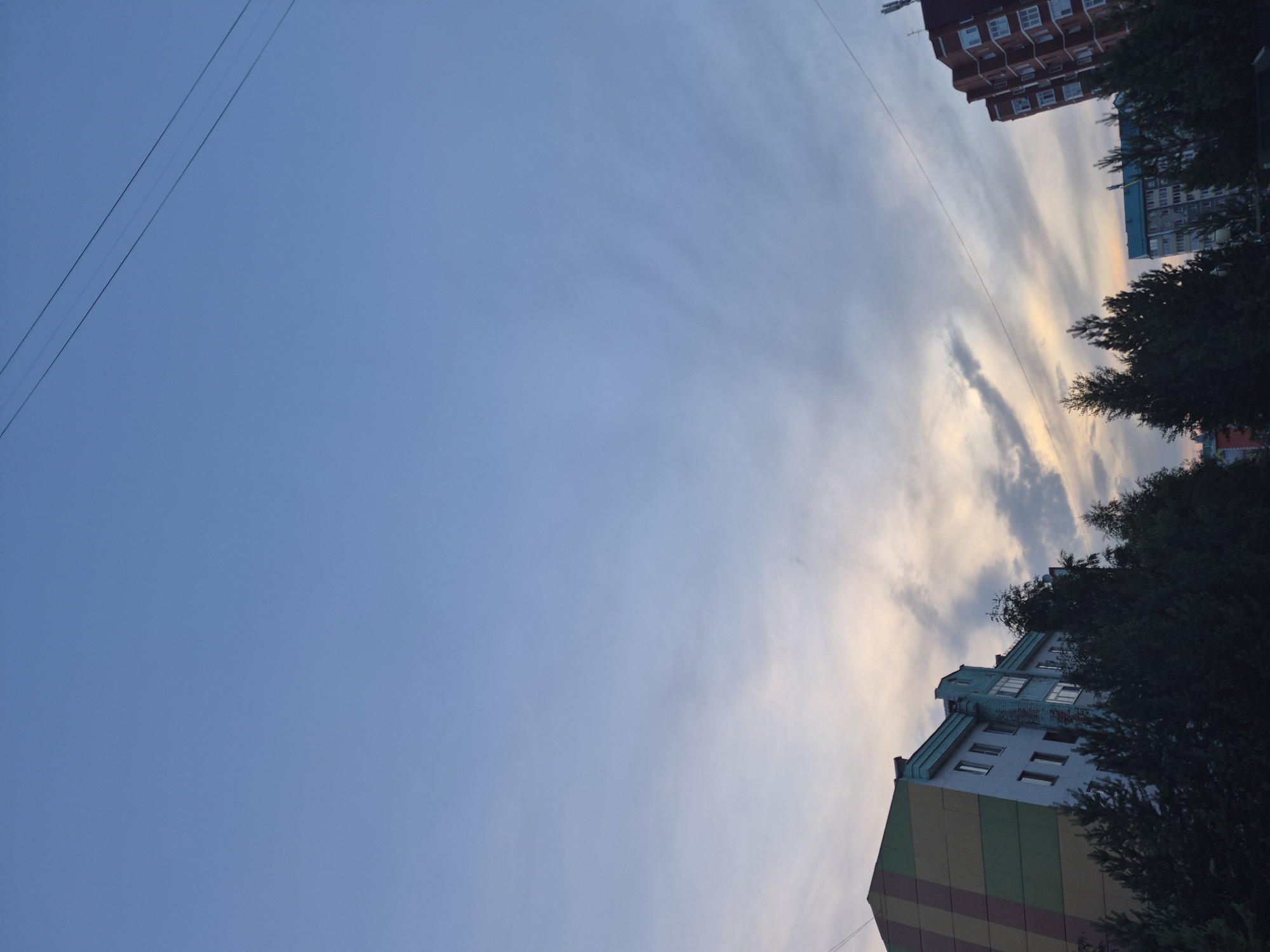 The image depicts a street view in the city of Nyagan, Russia, likely during the late afternoon or early evening, as indicated by the soft lighting and cloudy sky. In the foreground, several trees are visible, some of which appear to be coniferous. Behind the trees, two multi-story buildings are present. The building on the left has a distinct facade with horizontal bands of green, yellow, and brown. The building on the right is a red brick structure with a unique architectural design featuring stepped levels and numerous windows. A power line stretches across the upper portion of the frame, bisecting the sky. The sky itself is a pale blue, transitioning to lighter hues towards the horizon, with wisps of clouds catching the fading sunlight. There are no people or vehicles visible in the scene. The overall impression is one of a quiet urban setting under a transitional sky.
