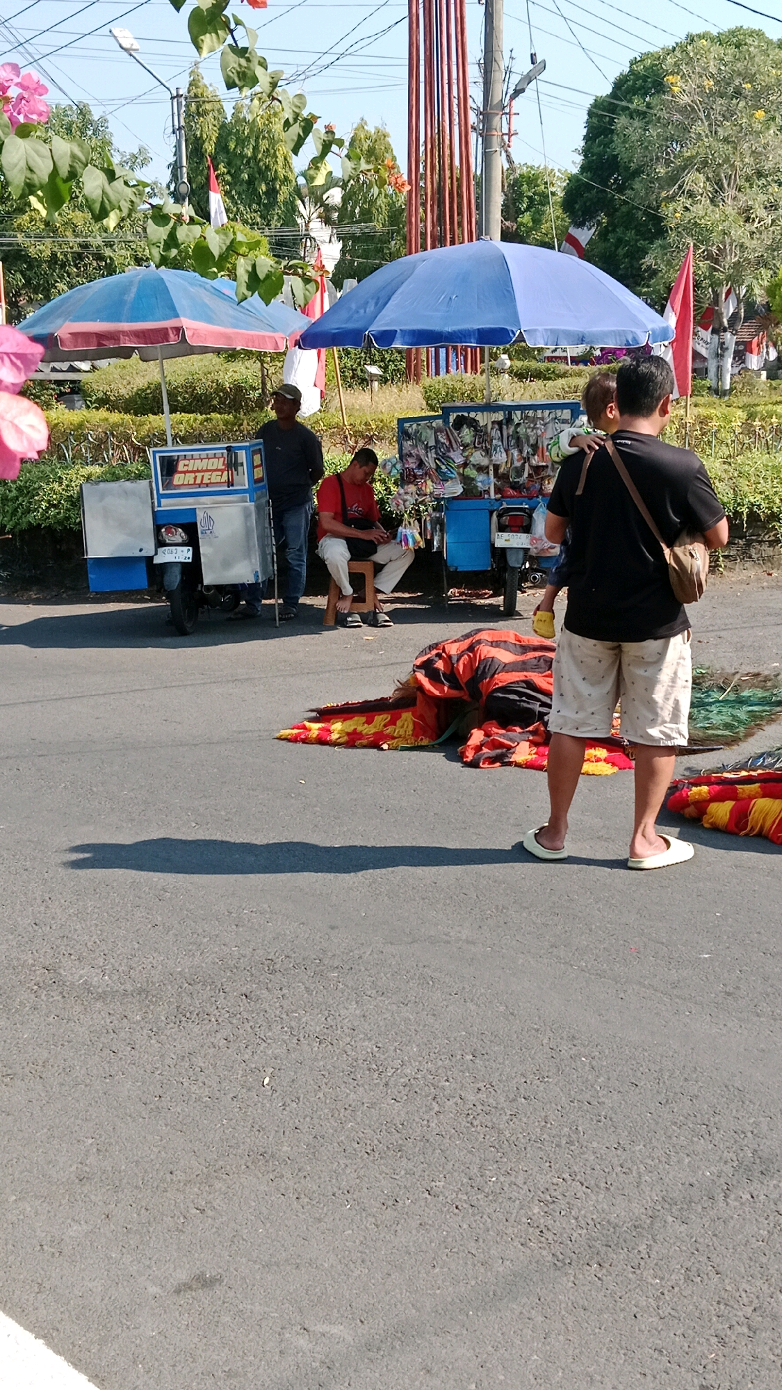 The image captures an outdoor scene in Magetan, Indonesia, under a bright, clear sky. In the foreground, the asphalt road dominates the lower half of the frame, with a distinct shadow cast by a person standing on the right. On the road, a colorful textile, predominantly red and black with yellow fringe, lies spread out. To the left of this textile, a person wearing a black t-shirt and light-colored shorts stands facing away from the camera, holding a yellow object in their right hand and carrying a sling bag across their chest. To their left, and closer to the background, a person in a red shirt and white pants is seated on a stool, looking down at an object in their lap. Beside them, a blue and white food cart is parked, displaying various items on its shelves. Another similar cart is visible further to the left, covered by a blue and red umbrella. In the background, lush green trees and foliage are abundant, with a tall pole structure featuring red vertical elements. Indonesian flags, red and white, are visible, adding a touch of national identity to the scene. The overall impression is of a street-side vending area, possibly a market or a roadside attraction, on a sunny day in Magetan.