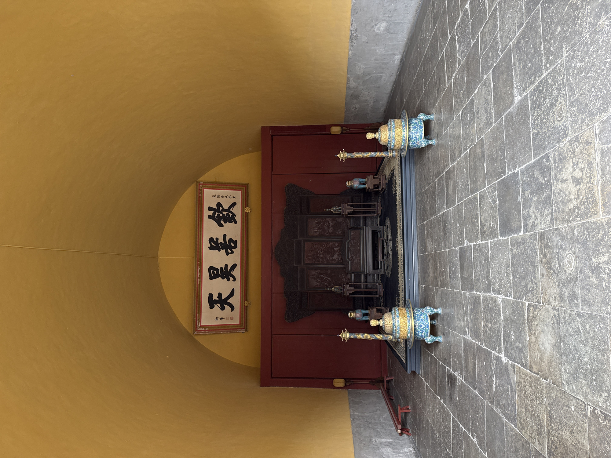The scene depicts a ceremonial area within a building in Dongcheng District, China. The space features an arched, gold-yellow ceiling and upper walls. The lower walls are grey and roughly textured. The floor consists of large, uneven grey stone tiles.
At the rear, a dark red paneled wall serves as a backdrop, slightly recessed from the main floor level. Above this wall, a large, rectangular framed plaque displays three prominent black Chinese characters, likely "敬天勤民" (Revere Heaven and Diligently Govern the People), flanked by smaller inscriptions, on a light background with a red border.
Centered on a raised, dark platform in front of the red wall is an elaborate dark wooden throne or ceremonial chair with a high, intricately carved backrest and armrests. A dark blue patterned rug covers the platform beneath the throne. To the left and right of the throne, on the same platform, are smaller dark wooden stands, each supporting a small decorative statue: a seated figure on the left and a quadrupedal animal, possibly a foo dog, on the right. A low, dark wooden bench or footrest is positioned in front of the throne.
On the main tiled floor, to the left and right of the raised platform, are two large, lidded, ornate censers. These censers are primarily turquoise, gold, and blue, with intricate patterns, and stand on three legs. Each censer has a tall, slender, golden-topped decorative pole or finial extending upwards from its side.
To the far left of the scene, near the grey wall, a simple, low red wooden bench is present. No people are visible in the scene.