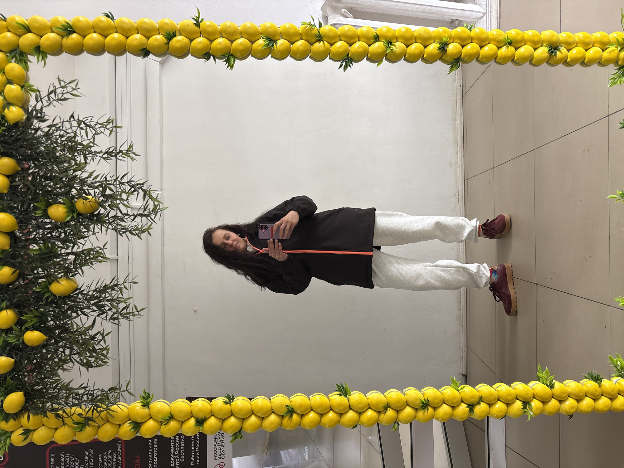 A woman is taking a selfie in a mirror decorated with artificial lemons and greenery. The mirror is likely indoors, possibly in a commercial or public space. The woman is holding her phone in her right hand and looking at the screen. She is wearing a dark jacket with an orange accent line, grey sweatpants, and brown sneakers. The background is a tiled floor and a wall. There is some text visible near the bottom left corner of the image, partially obscured by the lemon decoration. The presence of Russian text suggests the location is likely in Russia. Given the OCR text containing "России" (Russia), it is likely that the location indicated is indeed in Russia. Specific location details are not present in the provided context.