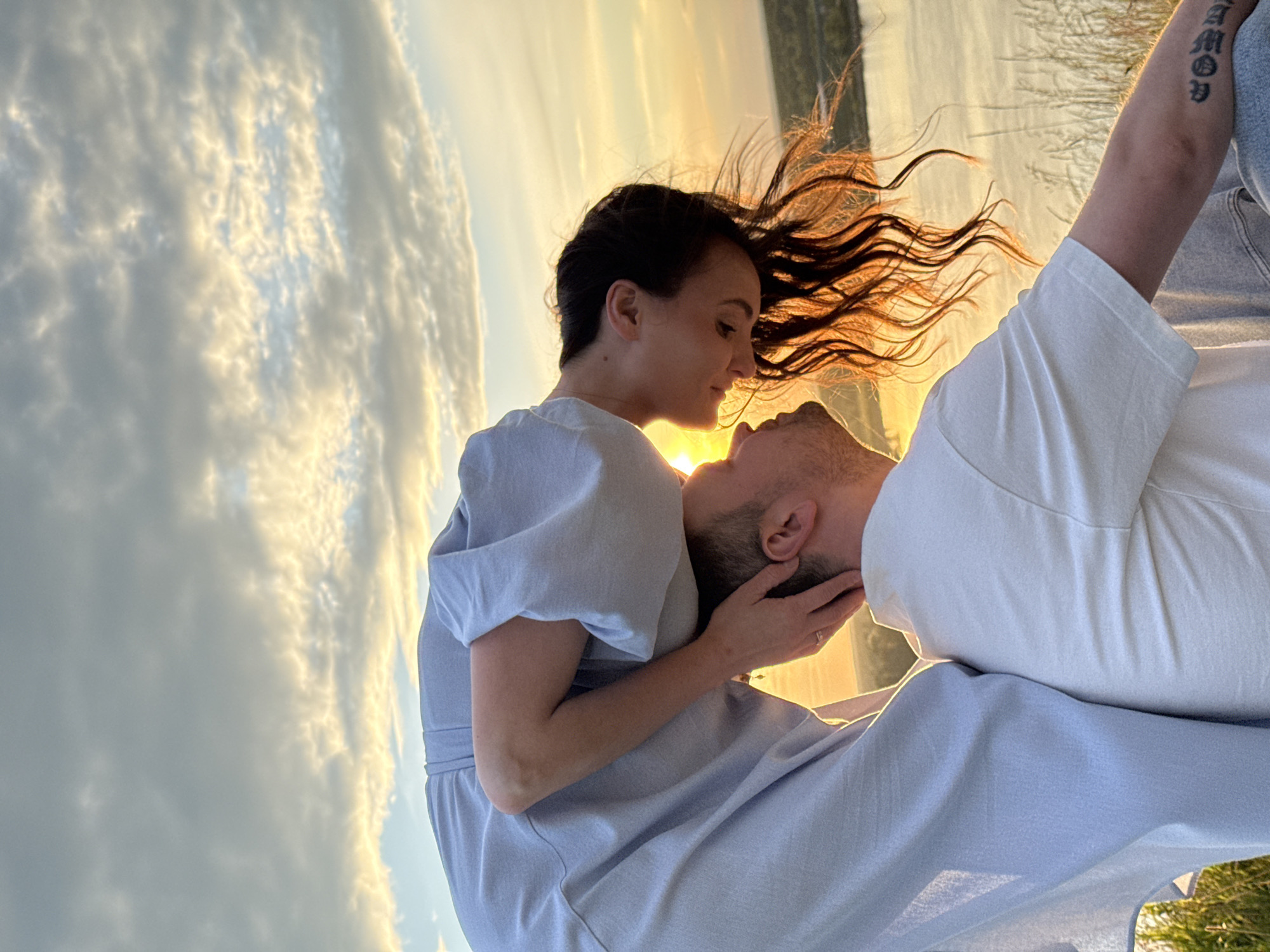 The image depicts a romantic moment between a couple against a backdrop of a sunset and sky. A woman, with her dark hair flowing in the wind, is leaning towards a man. She is wearing a light blue dress, and he is in a white t-shirt. The setting appears to be outdoors, possibly near a body of water, with reeds visible in the background and the golden hues of sunset illuminating the scene. The sky is filled with dramatic, wispy clouds catching the sunlight, suggesting either late afternoon or early evening. The man has a tattoo on his arm with the text "AMOD" visible. The overall mood is intimate and tender, with the couple seemingly lost in their own world as the sun sets. The image is captured in a vertical orientation.