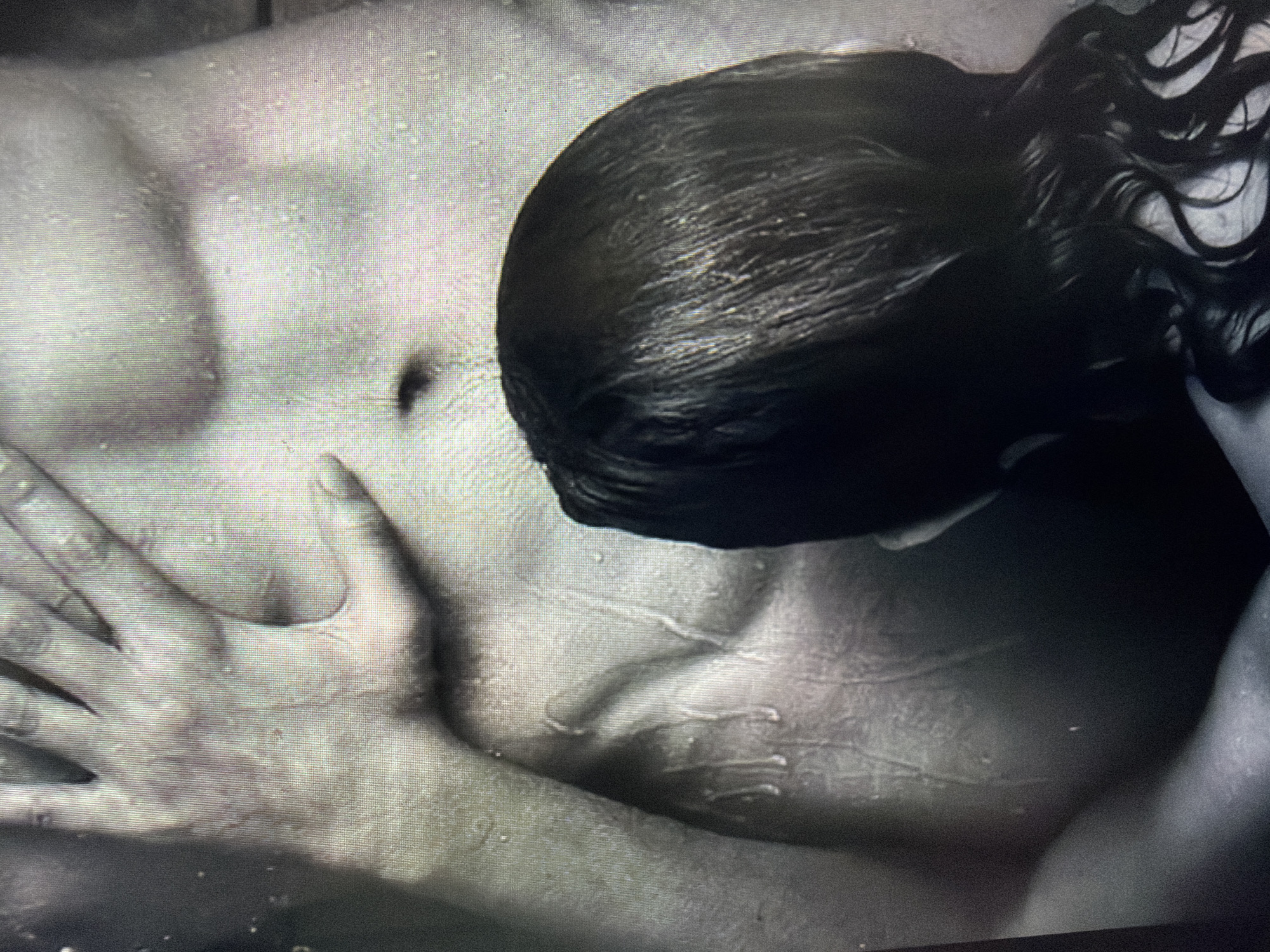 The media file is a close-up, grayscale image focusing on the torso and arm of a person. The subject appears to be lying down, with their head, covered by dark, wet hair, resting on their chest or shoulder. Water droplets are visible on the skin, suggesting a wet environment such as a shower or pool. The lighting is dramatic, creating strong shadows that accentuate the musculature of the abdomen and chest. A hand with visible fingers is positioned across the abdomen, with its palm and fingers gently touching the skin. The overall mood is intimate and perhaps contemplative, with the water and shadows contributing to a sense of vulnerability or relaxation. There is no visible text.
