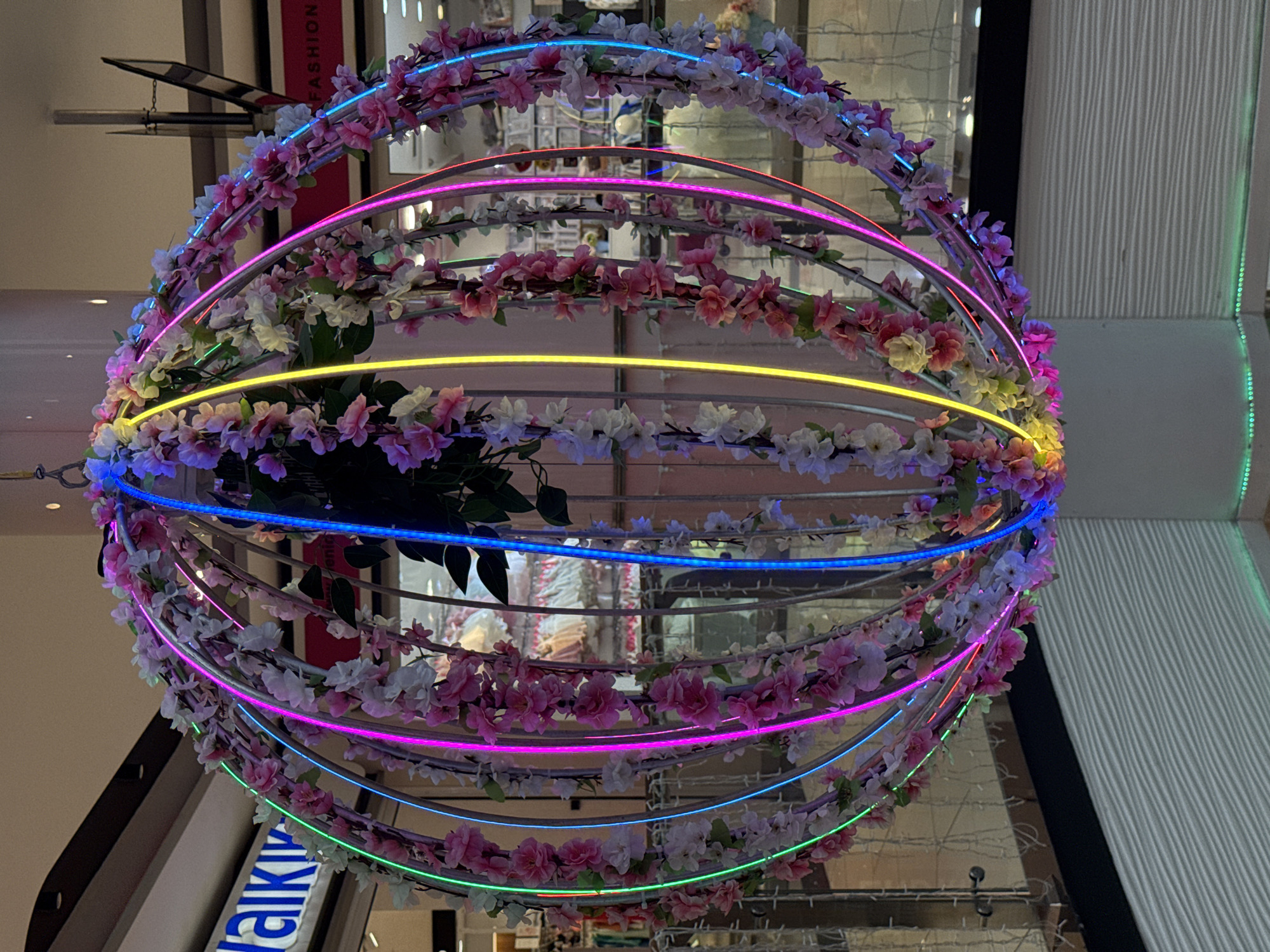 The image shows a large, spherical floral decoration hanging from the ceiling of an indoor space, likely a shopping mall or public building. The sphere is constructed of metal hoops adorned with artificial pink and white flowers and greenery. Integrated into the structure are colorful LED lights that appear to be cycling through various colors, including green, blue, yellow, and pink.
In the background, there are signs that appear to be for retail stores. One sign clearly visible reads "FASHION" in red lettering. Another sign to the left has white letters on a blue background, with the visible portion reading "AIKI". The overall environment suggests a commercial setting.
The image does not contain any people or animals. The lighting is a mix of ambient interior light and the vibrant glow from the LED decorations. The perspective is from below, looking up at the suspended ornament. There is no specific indication that this scene is located in Baku, Azerbaijan.