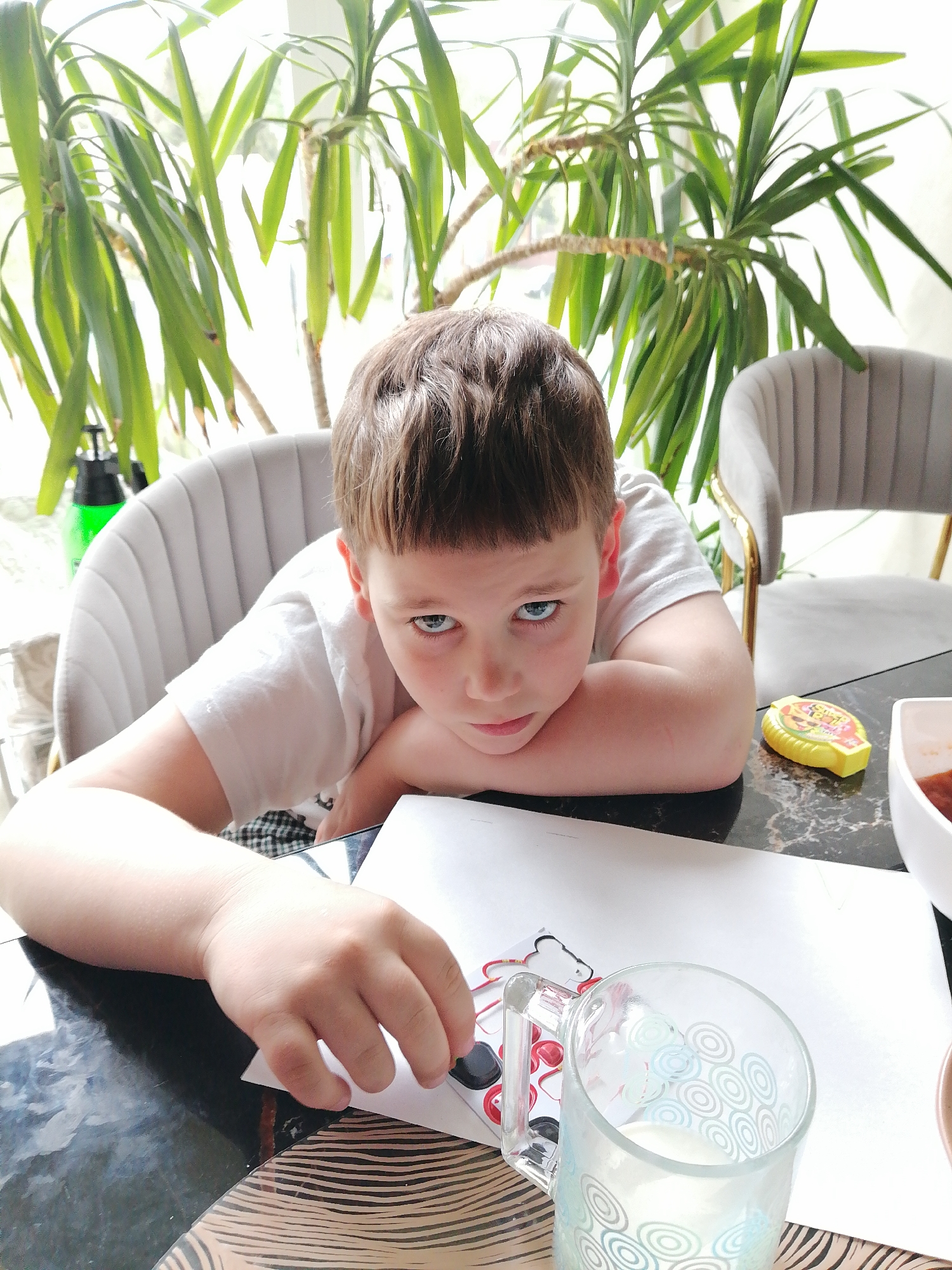 A young boy with light brown hair and blue eyes sits at a table. He is wearing a white t-shirt. His head is resting on his arms. On the table is a sheet of white paper, a glass of milk, a yellow container, and a white bowl. A green bottle is visible in the background. Two chairs are behind him, and a leafy plant fills the background. There is no verifiable information about the location of the image. Trubacheyevka is a village in Russia.