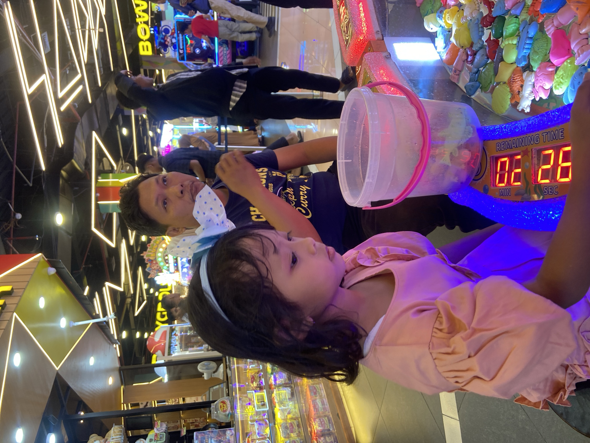 In a brightly lit indoor arcade in Sidoarjo, Indonesia, a young girl with a blue headband and a pink puff-sleeved dress is focused on an arcade game. She is positioned in the foreground, to the left of the frame, her gaze directed downwards towards the game's playfield. Beside her, on the right, a clear plastic bucket filled with water sits on a colorful assortment of plastic sea creatures, which are part of the game's mechanics. A digital timer on the game display shows "MIN 06" and "SEC 26" beneath the text "REMAINING TIME". Behind the girl, a man wearing a dark blue t-shirt with yellow text and a white polka dot mask pulled down to his chin, watches the game. The background reveals a lively arcade environment with other patrons, game machines, and overhead fluorescent lighting.