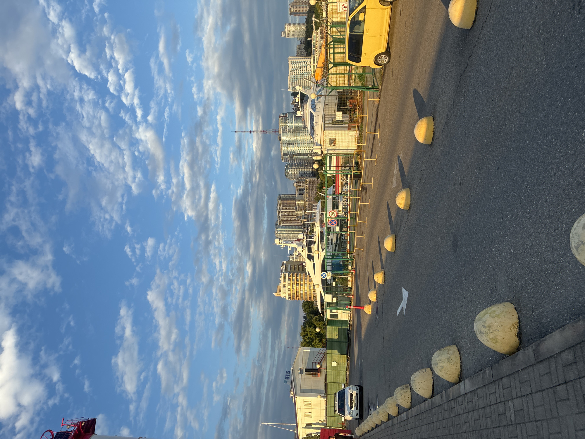 The image shows a street scene in the city of Sochi, Russia, likely near a harbor or marina. The sky is blue with scattered white clouds, and the sun is casting long shadows, suggesting it is either early morning or late afternoon.
In the foreground, a paved road is lined with hemispherical concrete bollards. A white car is parked on the left side of the road, and a yellow car is parked on the right. Further down the road, there is a building with a red and white lighthouse structure attached to its left side. A green fence runs along the side of the road, separating it from a waterfront area with several yachts docked. In the background, modern high-rise buildings are visible, characteristic of a city skyline. The overall impression is one of an urban coastal environment under a pleasant sky.