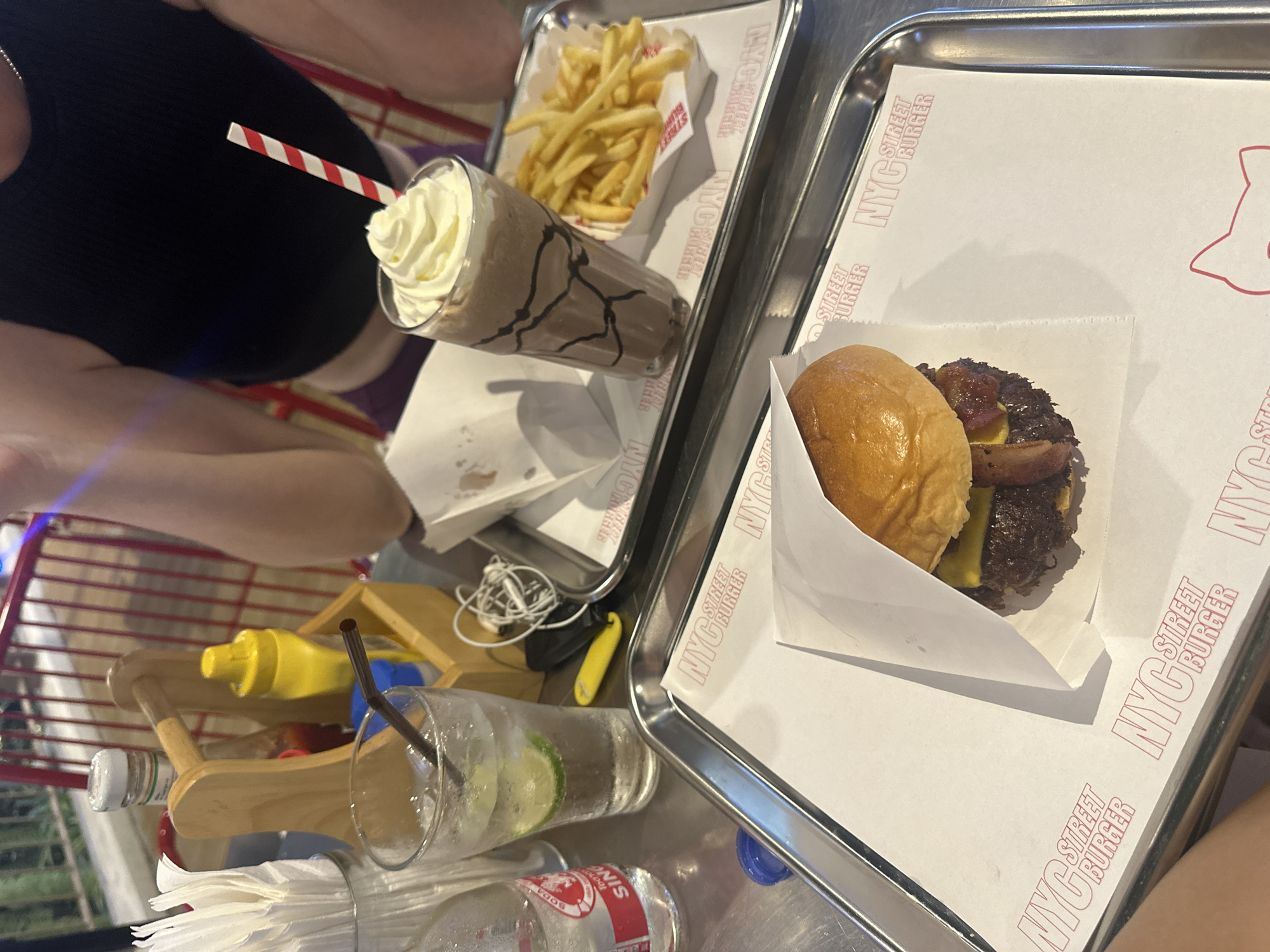 The image shows a meal at what appears to be "NYC Street Burger". There are two metal trays containing food. One tray has a burger wrapped in white paper with "NYC Street Burger" printed on it. The other tray has a milkshake with whipped cream and a striped straw, and a portion of french fries. There is also a glass with a straw and lime slices, along with bottles of soda or other condiments on the table. Part of a person is visible, wearing a dark-colored top. The table appears to be inside or in an outdoor seating area with red metal bars of chairs around. The environment suggests a casual dining setting in the city of บ้านบางรักษ์, Thailand.