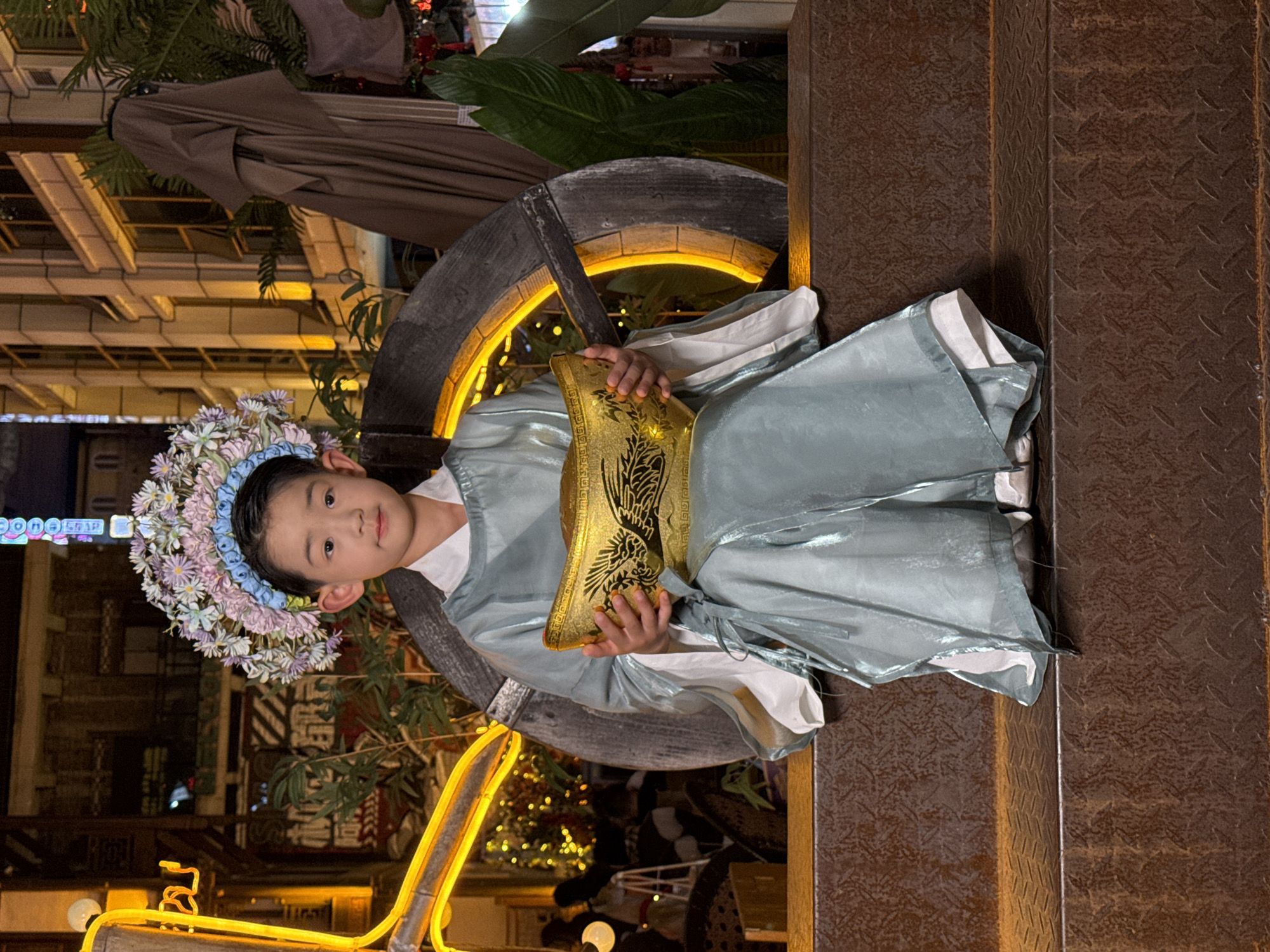 A young East Asian child, appearing to be a boy, is seated and looking directly at the viewer in a brightly lit, festive nighttime urban setting. He is wearing an elaborate light blue-green traditional-style outfit with white undergarments, and a large, colorful floral wreath or crown adorns his head, featuring shades of pink, blue, and white flowers. The child holds a golden, ornate rectangular clutch or small bag with black decorative patterns, possibly depicting a phoenix or similar mythological creature, in front of him. His expression is calm and composed.
The immediate background behind the child features a large, dark wooden wheel-like structure with an illuminated inner rim, casting a warm yellow glow. To the child's right, a textured metal surface with a diamond plate pattern, possibly a wall or pillar, is visible. The broader background shows blurry buildings with illuminated windows and green foliage, suggesting an urban street or plaza at night. A bright yellow neon-like light fixture is partly visible in the lower left foreground.
The scene conveys a festive and decorative ambiance, likely an evening photo opportunity in a commercial or entertainment district. The lighting suggests it is nighttime. Partial, blurry Chinese text is visible on signs in the distant background, consistent with the Yuzhong District, China location context.