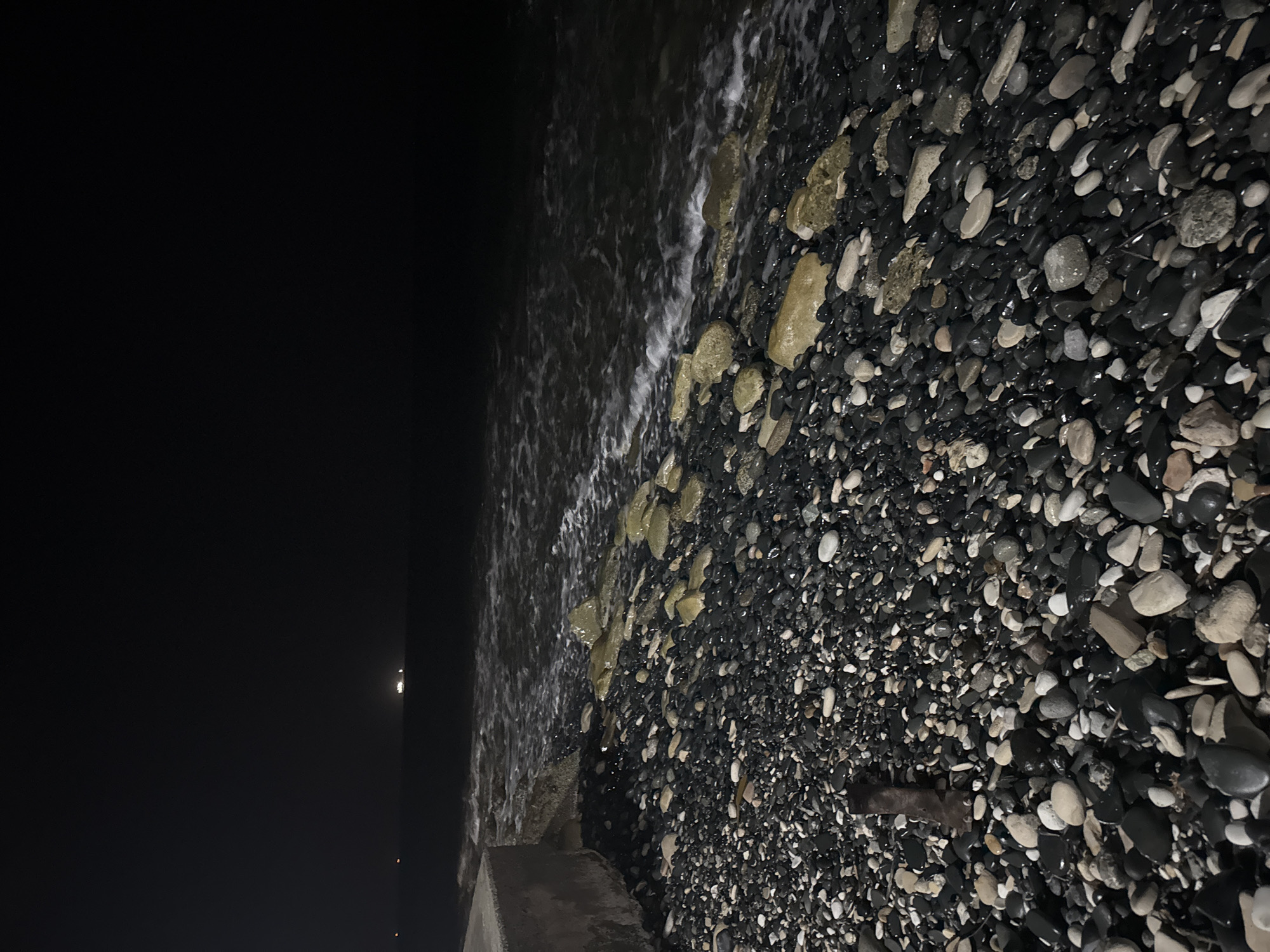 The image shows a pebble beach at night in the Agios Tychona Community, Cyprus. The foreground is dominated by a dense scattering of various sized, dark and light colored pebbles, some wet and reflective, others dry. Several larger, lighter colored rocks are interspersed among the pebbles closer to the water's edge. To the left, a portion of a concrete structure, possibly a breakwater or pier, is visible. The sea is to the right and in the background, with dark, choppy water and white foam from gentle waves breaking on the shore. In the far distance, across the dark expanse of the sea, a few faint lights are visible on the horizon, suggesting distant lights or possibly a ship. The sky is completely dark, indicating it is nighttime. There are no people or animals visible in the frame.