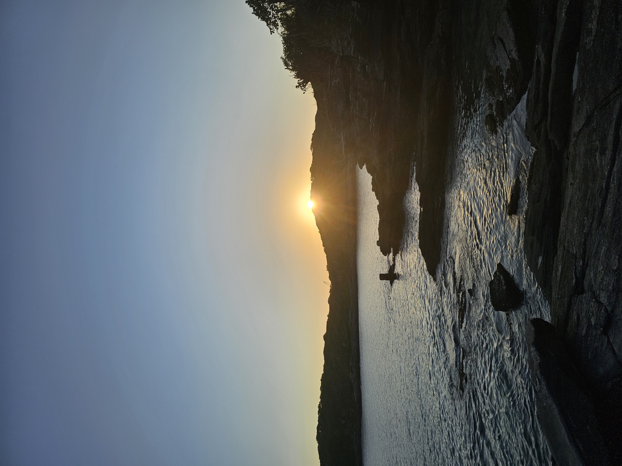 Here's a description of the image:
The image depicts a body of water, possibly the ocean or a bay, with land masses rising on either side. The sun is setting, with the sun's position appears to be directly behind the land in the middle of the image, indicating a horizon line. The lighting is generally dark and moody.
On the right side of the body of water is a large rock or cliff face, which is in shadow and textured with visible rock formations. Some water can be seen streaming down the right side of the frame. On the left side, there is a more gradual slope of land that is silhouetted against the setting sun.
In the body of water, there's what appears to be a person or small object centered in the middle of the water. The water itself is slightly turbulent with ripples.
Considering the location given as Armação dos Búzios, Brazil, and the land and rock formations, this could be a view of a sunset along the coastline.