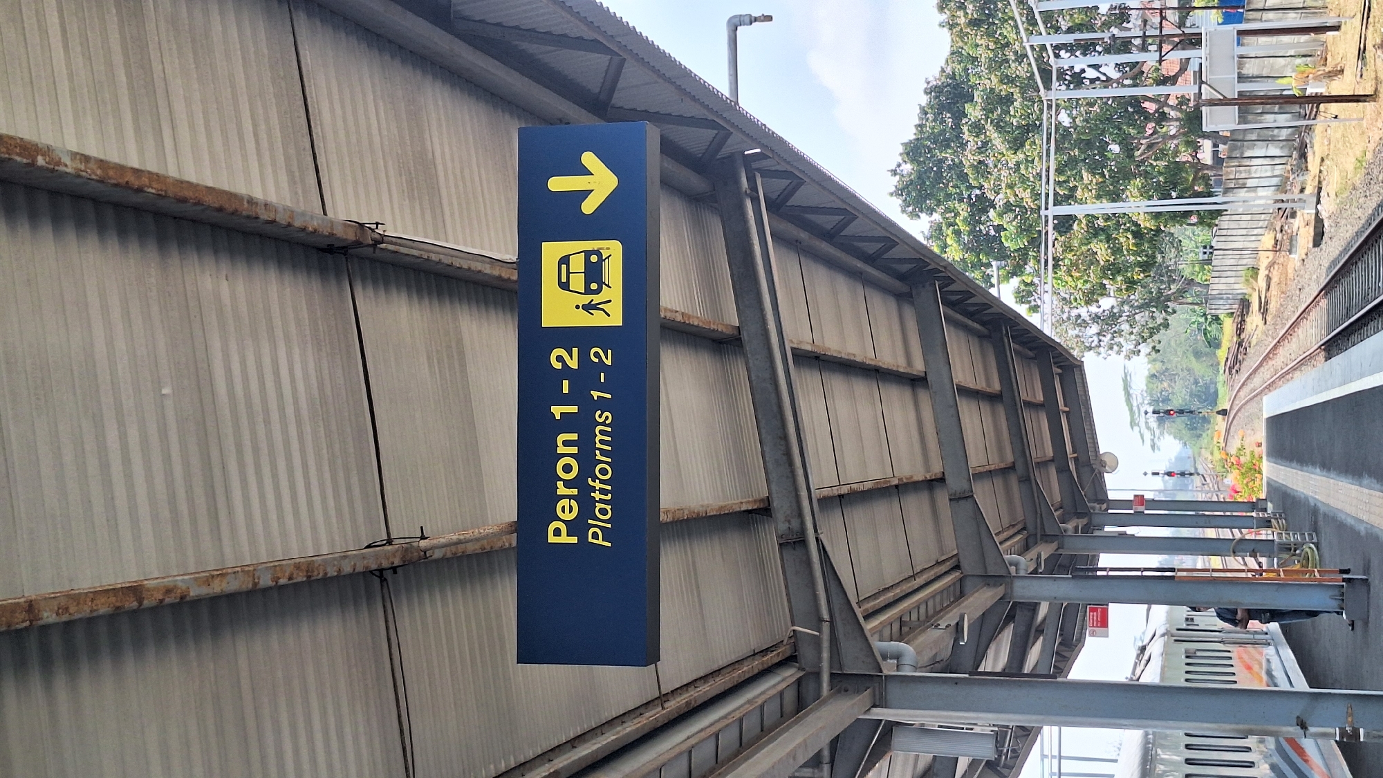 The image captures a sign at a train station in Kroya, Indonesia, indicating "Peron 1-2" and "Platforms 1-2" with directional icons. The sign is dark blue with yellow text and graphics, featuring a person walking towards a train and a downward arrow, suggesting the direction to platforms 1 and 2. The sign is suspended from a metal roof structure that appears weathered and shows signs of rust, supported by grey metal pillars.
The foreground shows a train on a track to the left, with its doors closed and windows visible. A person in dark clothing stands on the platform near the train, facing away from the camera. The platform itself is paved with a dark material, marked with yellow lines. To the right of the platform are train tracks curving into the distance.
The background reveals a bright, overcast sky and lush green trees and vegetation. The environment appears to be an outdoor train station, likely in a tropical or subtropical region, given the foliage. In the distance, traffic lights for the train are visible on the tracks. The overall impression is of an active, though perhaps somewhat aged, transportation hub.