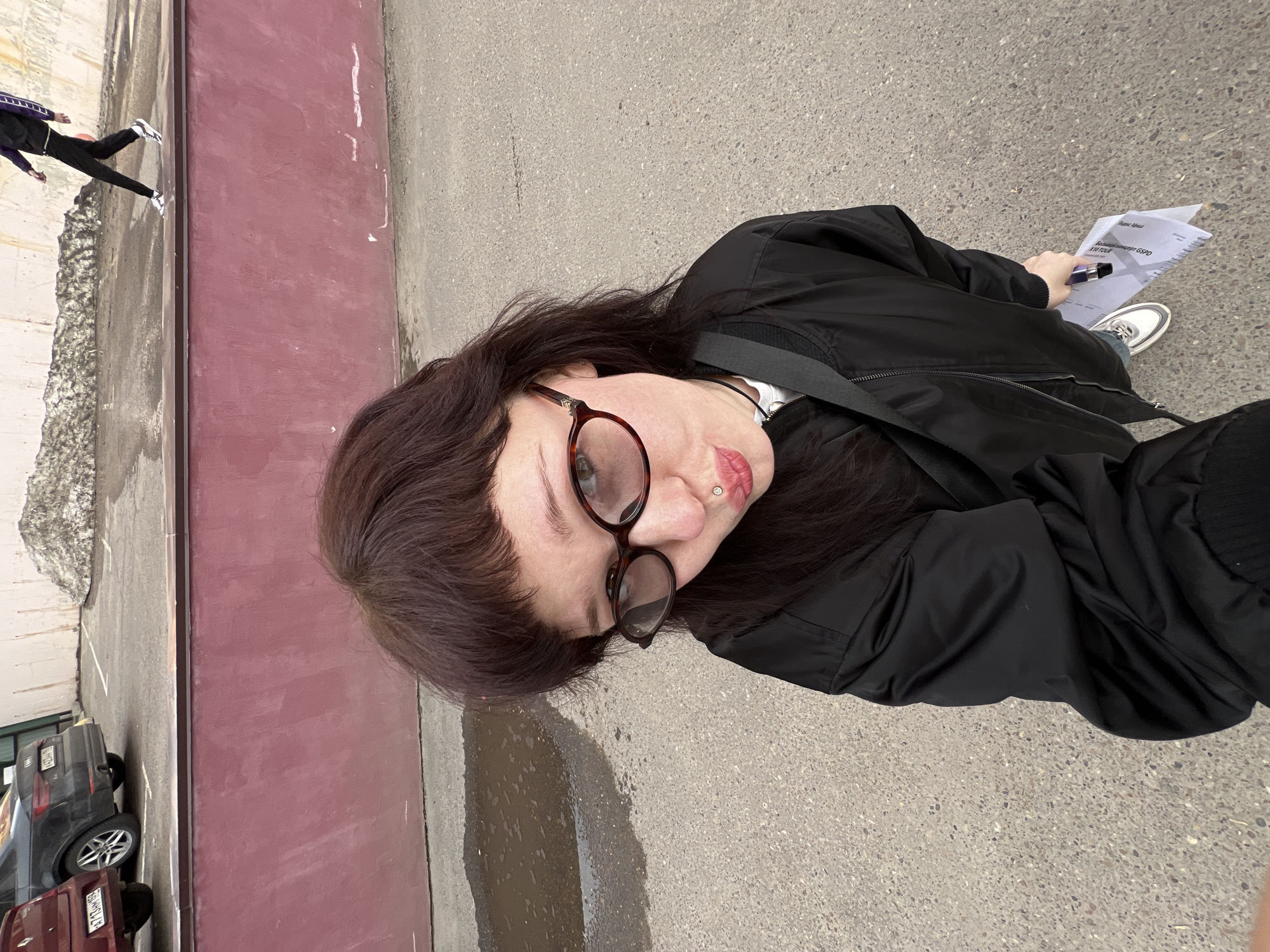 A woman is standing outside in what appears to be a city environment. The background includes a red brick wall, a paved area with a puddle, and glimpses of parked cars and buildings. She is wearing glasses and a black jacket and holding papers in one hand, while another person is walking by in the background. The location is Kazan, Russia.