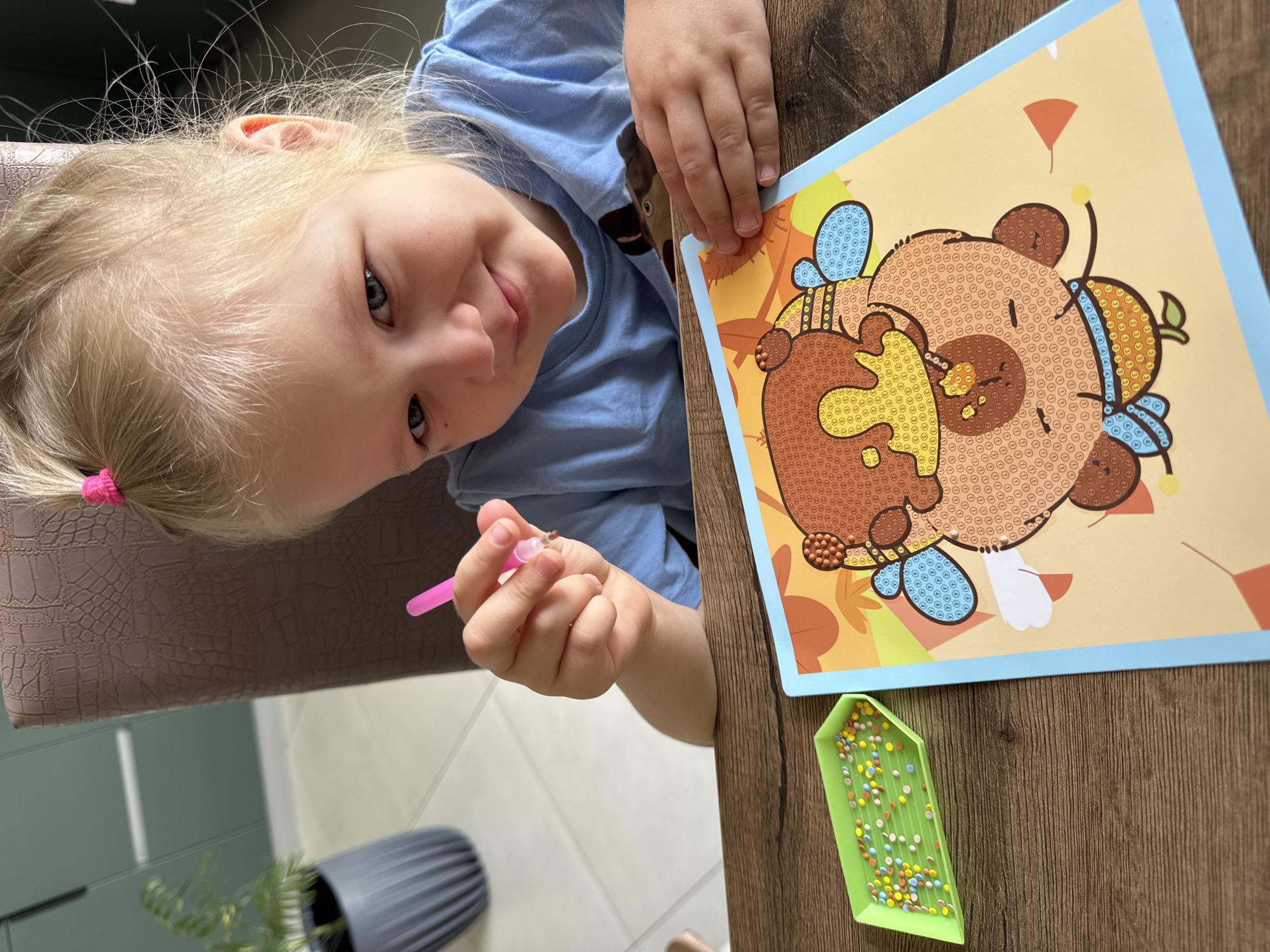 A young child with blonde hair and blue eyes is seated at a wooden table. The child is wearing a blue shirt and is holding a pink stylus in their right hand. The child is looking at the camera with a slight smile. On the table is a picture of a bear with a honey pot and a small green tray filled with colorful beads. The background includes a grey textured pot with a plant, and a brown chair. The location is likely indoors, possibly in a home. The image appears to be taken in the city of Ферма, Russia. The child appears to be engaged in a crafting activity.