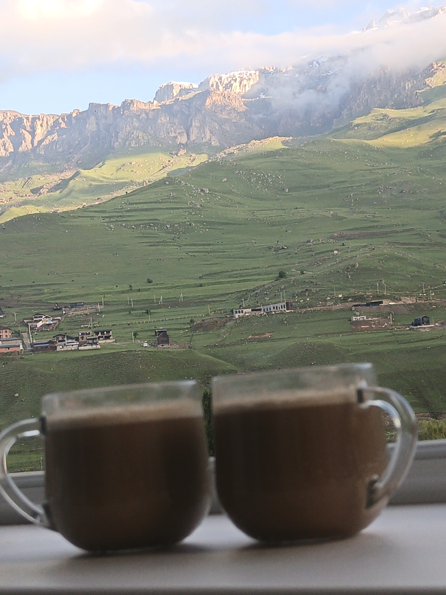 In the foreground, two clear glass mugs containing a light brown beverage, likely coffee, are positioned on a white surface. The mugs have distinct handles. Behind the mugs, a panoramic view unfolds of a verdant valley with rolling green hills, terraced fields, and scattered buildings that form a village. In the background, imposing mountains rise, their peaks partially obscured by soft, white clouds. The lighting suggests either early morning or late afternoon, casting a warm glow on the landscape. The village appears to be Verkhny Fiagdon, Russia, given the characteristic mountainous terrain and the style of the buildings visible.