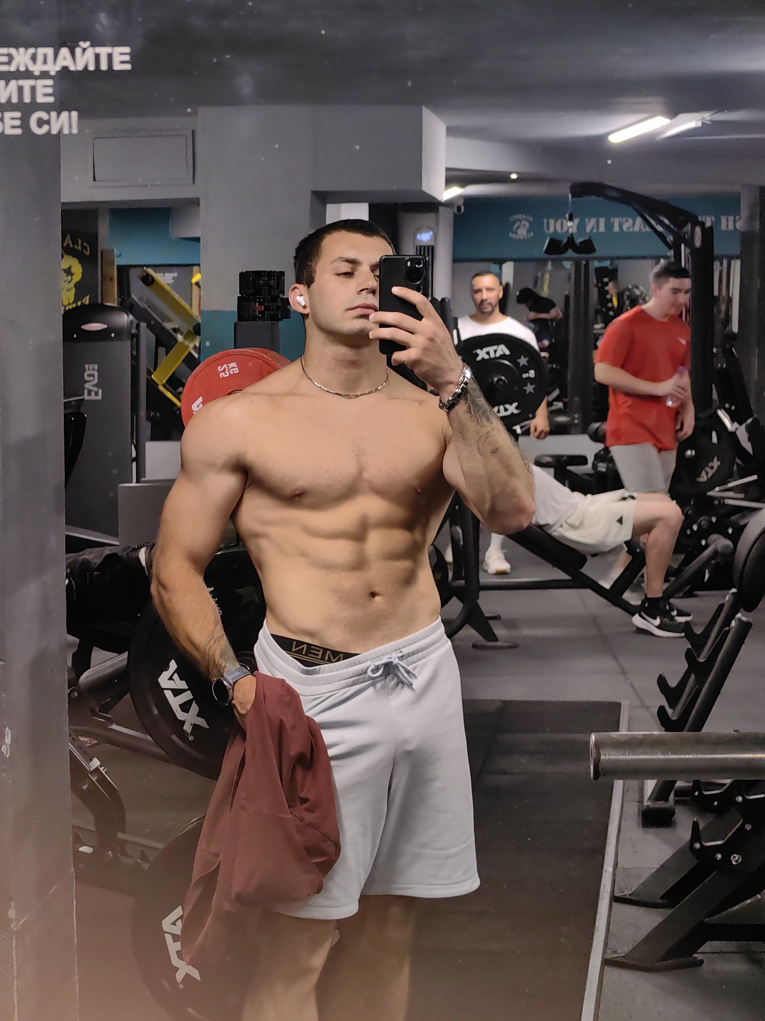 This is a selfie taken in a gym in Varna, Bulgaria. The main subject is a man with defined abdominal muscles, wearing light-colored shorts and a silver necklace. He is holding a phone in his right hand, taking the photo, and a dark red shirt in his left hand. He has white earbuds in his ears and a watch on his left wrist. The background shows a gym setting with weightlifting equipment, including barbells and weight plates. Other individuals are visible in the gym, one in a red shirt and another in a white t-shirt, seemingly engaged in their workouts. The lighting suggests it could be daytime or evening, typical for a gym environment. The overall mood appears to be one of fitness and self-care. Text is visible on a wall in the background, partially obscured by reflections, and also on the weight plates. The text on the wall, appearing as "ЕЖДАЙТЕ" and "ИТЕ Е СИ!", is in Cyrillic script. The weight plates have the text "XTA" visible.