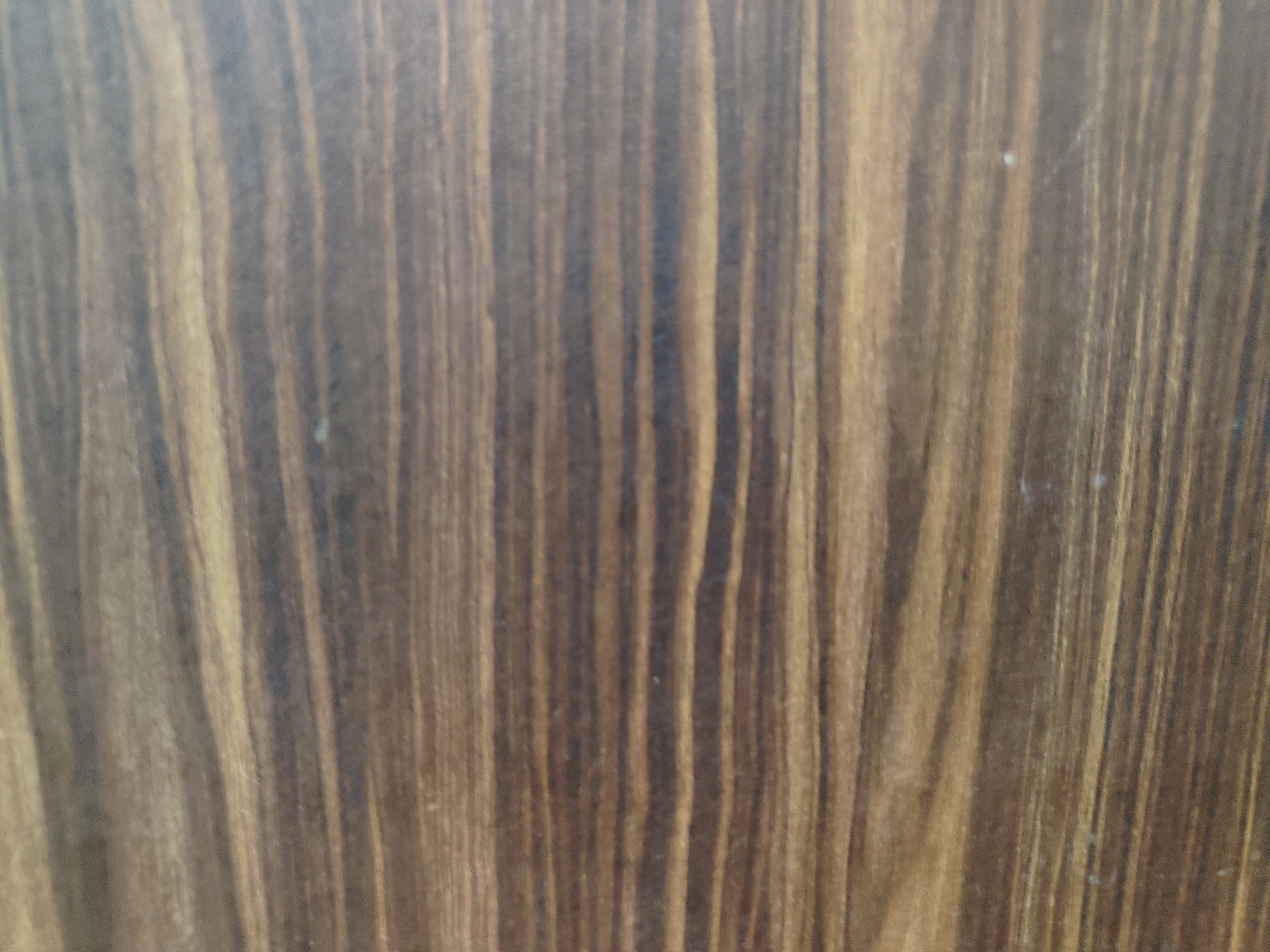 The image shows a close-up view of a wooden surface with a prominent grain pattern. The wood is a medium brown color with darker vertical streaks, suggesting it is a type of hardwood with a linear grain. The surface appears to be a flat panel, possibly a tabletop, a door, or a piece of furniture. There are no people, animals, or other objects visible in the image. The lighting is even, and the focus is sharp on the wood grain. The image does not contain any contextual clues that would indicate its location in Jamshedpur, India, or any other specific urban environment.