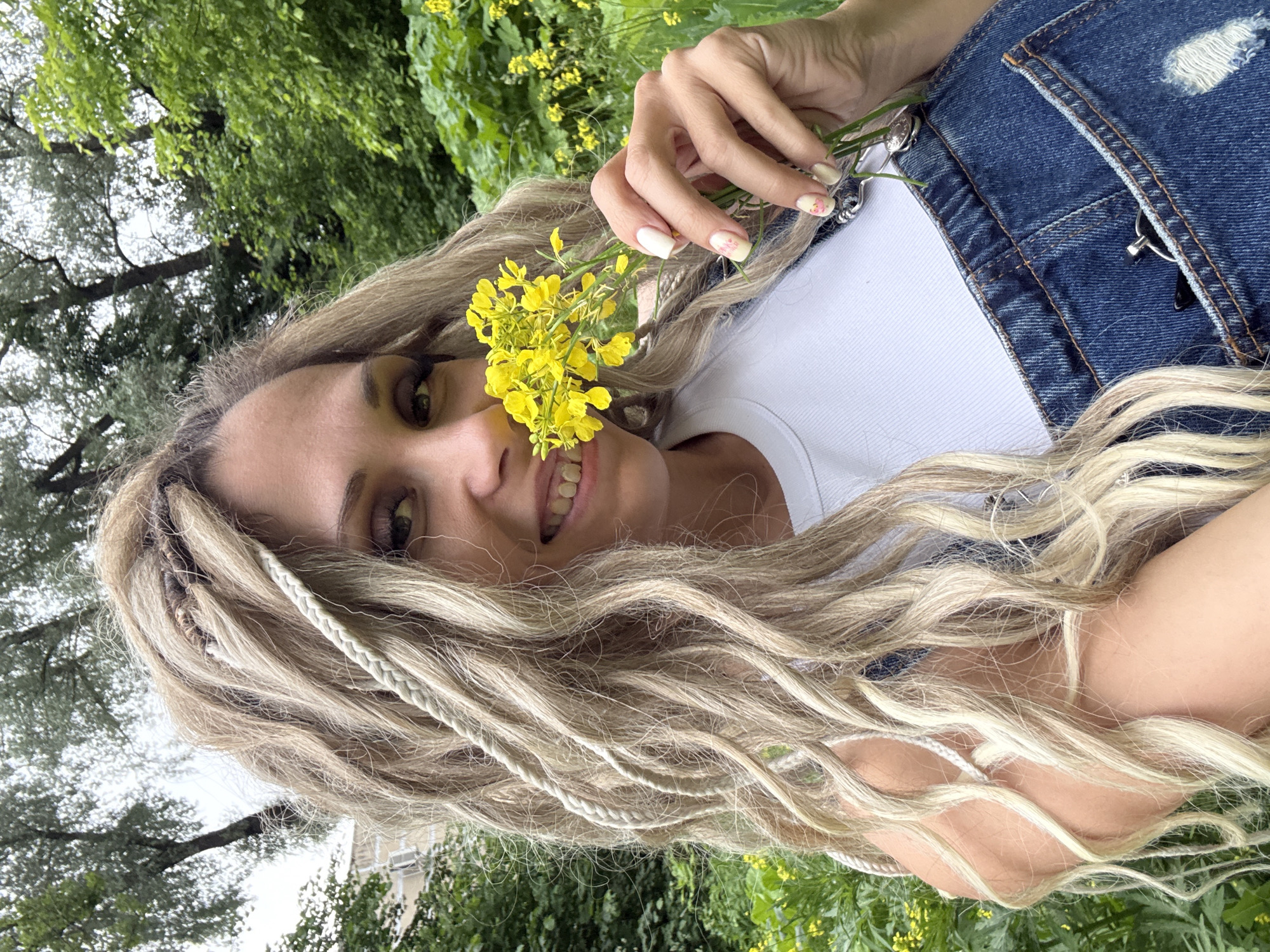 This is a close-up, outdoor, eye-level, medium shot of a young woman lying down amidst greenery. She has long, blonde dreadlocks that cascade around her face, some of which are braided. She is wearing a white t-shirt and denim overalls. Her right hand is raised, holding a small bunch of yellow wildflowers near her face, with the flowers partially obscuring her mouth. She is smiling, with her teeth visible. Her fingernails are painted white with a subtle floral design. The background consists of lush green foliage and trees, with hints of more yellow wildflowers scattered throughout. The lighting suggests it is daytime, possibly overcast due to the soft light and lack of harsh shadows. The overall mood is relaxed and cheerful, with the woman enjoying nature. There is no visible text in the image. The location context is Rusanovo, Russia.
