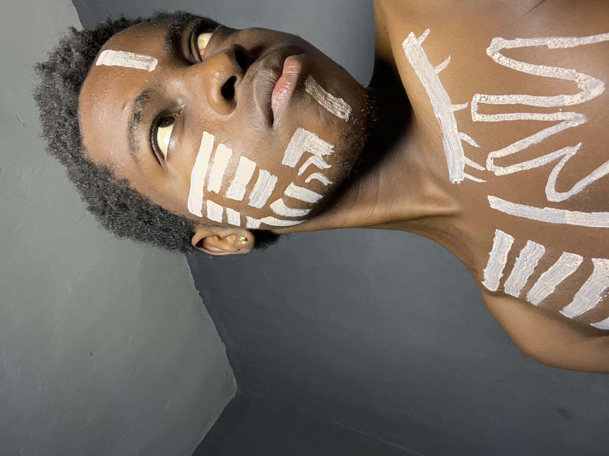 A young Black man with short, dark curly hair is depicted in a close-up shot from a low angle. He has white paint markings on his face and chest, consisting of vertical stripes and abstract shapes. His skin tone is dark brown, and he has a gold stud earring in his left ear. The background is a gray wall, and the image has been rotated, giving the impression that the man is lying down or looking upwards. The location of the image is not specified, but no information indicates that it is in Nkpor, Nigeria.