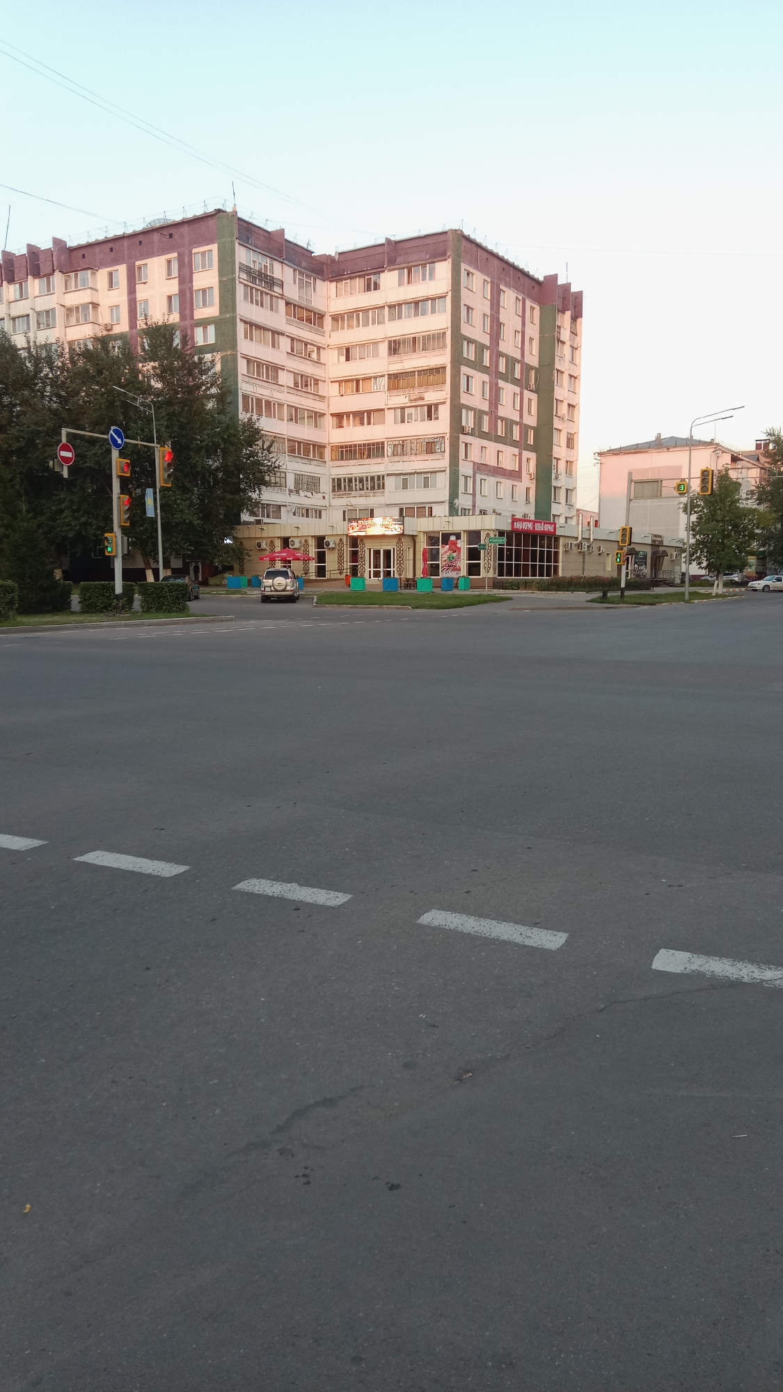The image depicts an intersection in the city of Petropavl, Kazakhstan, bathed in the soft light of either sunrise or sunset. Dominating the scene is a multi-story residential building with a distinct curved facade, its windows reflecting the sky. In front of this building, at street level, are storefronts, including one with red umbrellas and outdoor seating, suggesting a cafe or restaurant.
Several cars are visible on the road, including a white SUV parked near the storefronts. Traffic lights are present at the intersection, indicating controlled traffic flow. The road itself is asphalt with white dashed lines marking lanes. Trees and manicured bushes line the sidewalks, adding a touch of greenery to the urban landscape. The sky is a clear, pale blue, characteristic of a fine weather day. The overall impression is one of a typical urban environment captured during a transitional time of day.