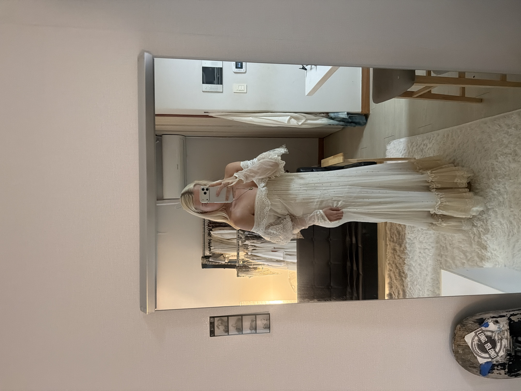 A person is seen in a mirror taking a selfie in what appears to be a fitting room or dressing area. The person is wearing a long, off-the-shoulder white dress with lace details. Behind the person, racks of clothing are visible, suggesting a boutique or store setting. The reflection in the mirror also shows parts of the room, including a shaggy white rug, furniture, and a wall with a light switch and other mounted fixtures. A skateboard with a "Long Island" sticker is placed on the corner shelf of the wall. The location in the city of Seoul, South Korea, cannot be determined from the given details of this image.