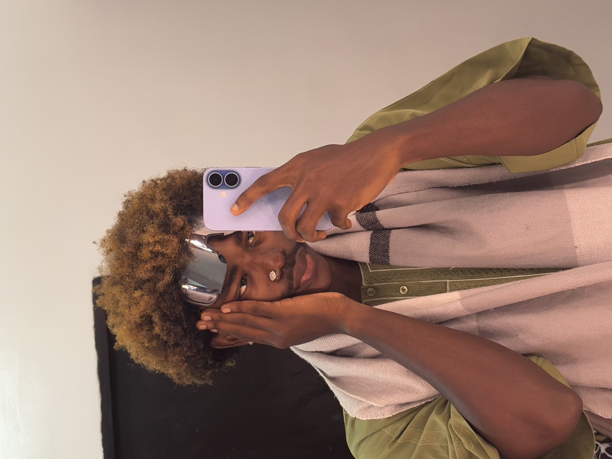 A close-up image depicts a dark-skinned individual, identified as male, situated indoors in Igboefon, Nigeria. The individual presents with brown, curly hair styled into an afro, a faint mustache, and a small, circular metallic nose piercing with a clear stone on the left nostril. Mirrored, reflective sunglasses are positioned on the forehead. The subject is attired in a green long-sleeved shirt featuring subtle patterned textures and a buttoned placket, overlaid with a light grey and white striped scarf draped around the neck and shoulders. The right hand holds a light purple smartphone, displaying a dual-camera system, directed towards the subject's face, indicative of a selfie capture. The left hand is raised, with fingers lightly touching the left temple area. The background consists of a plain light beige wall on the right and a dark, likely black, surface or backdrop on the left.