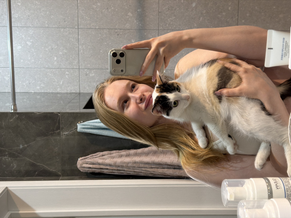 A young female, appearing to be in a bathroom in the city of Rossiyskiy, Russia, is captured in a mirror selfie. She has long blonde hair and is wearing a light-colored garment, possibly a camisole or bra. In her right hand, she holds a silver-colored smartphone with a triple camera array, pointed towards the mirror to take the photograph. With both arms, she cradles a calico cat, predominantly white with black and orange/brown patches and green eyes, which is looking downwards. The female's lips are slightly pursed, and she glances towards her right. Her fingernails are painted a light pink. In the foreground, on a surface below the mirror, several toiletries are visible, including two white pump bottles, one labeled "CLEAN" with Cyrillic text, and a white tube. The bathroom features dark grey marble-patterned tiles on the left wall and lighter grey speckled square tiles on the right wall. A silver towel rail is mounted horizontally above the lighter tiles. Two silver hooks on the dark grey wall support a textured light brown towel and a light blue towel. A white door frame is partially visible on the far left.