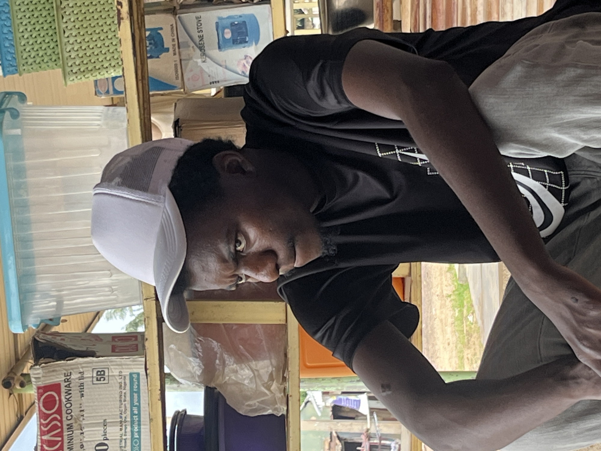 This is a medium shot photo of a person with dark skin wearing a black t-shirt and a white trucker hat. The person is looking down and appears to be focused on something outside the frame. They have dark, curly hair and a goatee. The background is cluttered with various objects, including plastic containers, boxes, and what appears to be shelving. Some of the boxes have text, with one clearly visible stating "CASSO ALUMINIUM COOKWARE" and another reading "KEROSENE STOVE MADE IN CHINA". There is also text about "product all year round". The overall setting appears to be a storage area or a market stall. The lighting suggests it could be daytime, possibly outdoors or in a well-lit indoor space with natural light coming in from the background. The person's expression is intense, but it's unclear what they are doing or if they are interacting with anyone. The image is oriented sideways.