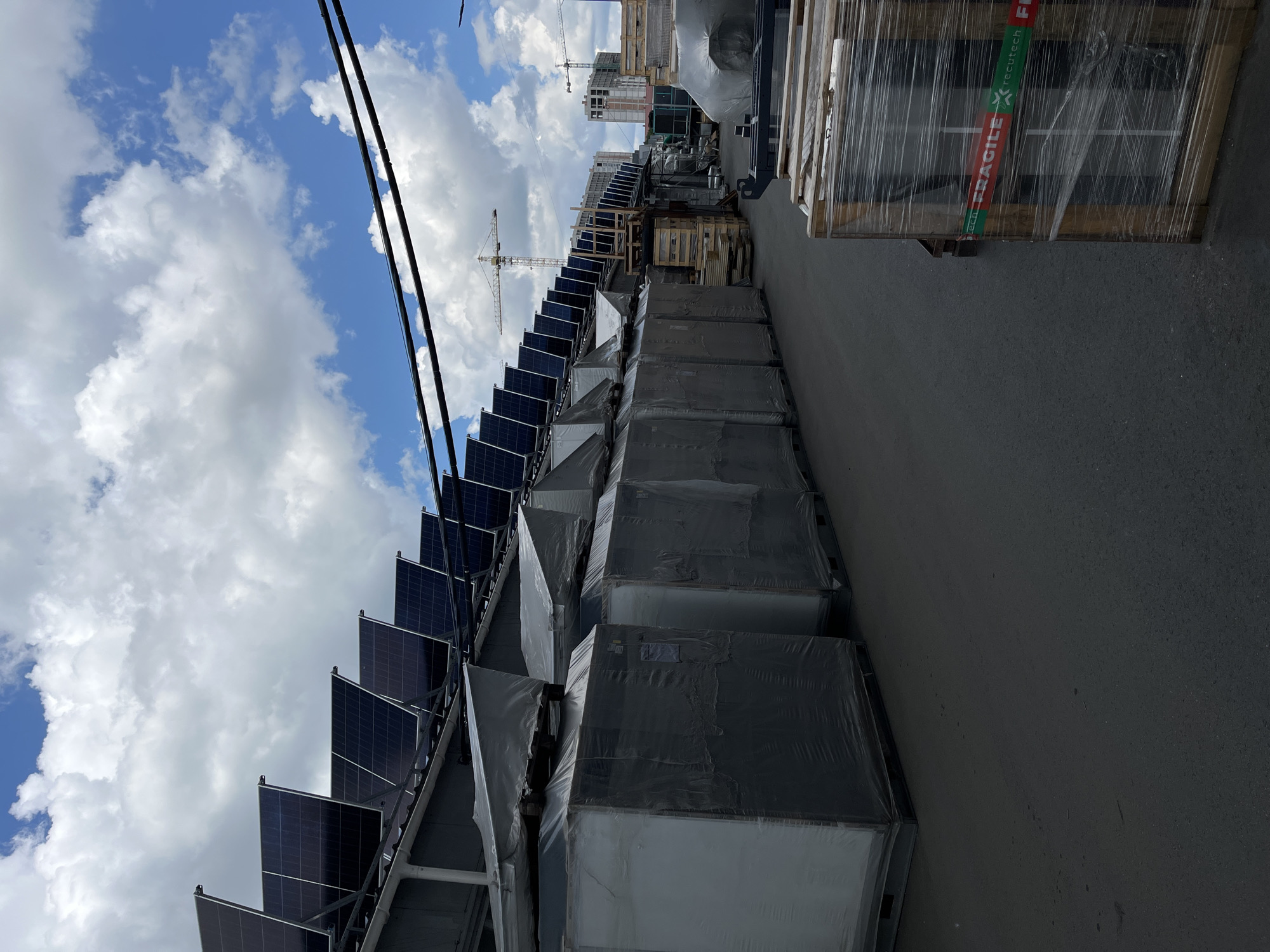 The image depicts an outdoor industrial or storage area with a row of large, wrapped items on pallets. The items appear to be rectangular and are covered in translucent plastic wrap. Above these, a series of solar panels are mounted on a tiered structure, angled towards the sky. In the background, construction cranes and buildings are visible, suggesting an urban or suburban environment. The sky is cloudy with patches of blue. The ground is paved. A pallet with a wooden crate containing what appears to be glass panes, wrapped in plastic and secured with tape labeled "FRAGILE" and "recutech", is in the foreground on the right. The scene is likely in the city of Kyiv, Ukraine, given the context.
