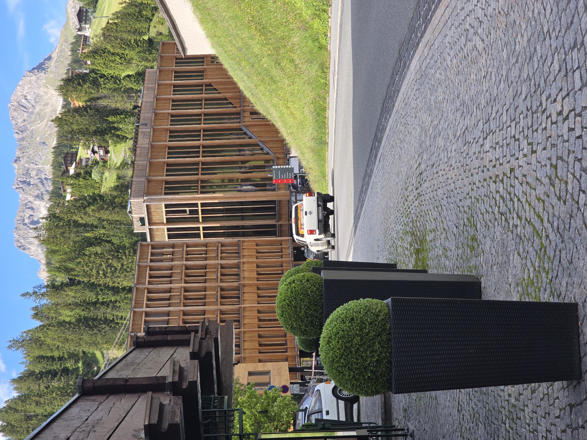 The image depicts a street scene in Lech, Austria, characterized by modern wooden architecture and a mountainous backdrop. In the foreground, several large black planters with perfectly spherical green shrubs are positioned along a cobblestone sidewalk. To the left, a white car is parked, and a portion of a building with dark wooden beams is visible.
Further into the scene, a white pickup truck drives away from the camera on an asphalt road. Behind the truck, a prominent building with a facade of vertical wooden slats and large glass windows stands out. To the right of this building, a grassy hill slopes upwards. The background is dominated by a dense forest of pine trees, with scattered houses and chalets nestled on the hillsides. Towering in the distance are rugged, rocky mountains under a clear blue sky. The overall impression is one of a modern resort town in a mountainous alpine setting.