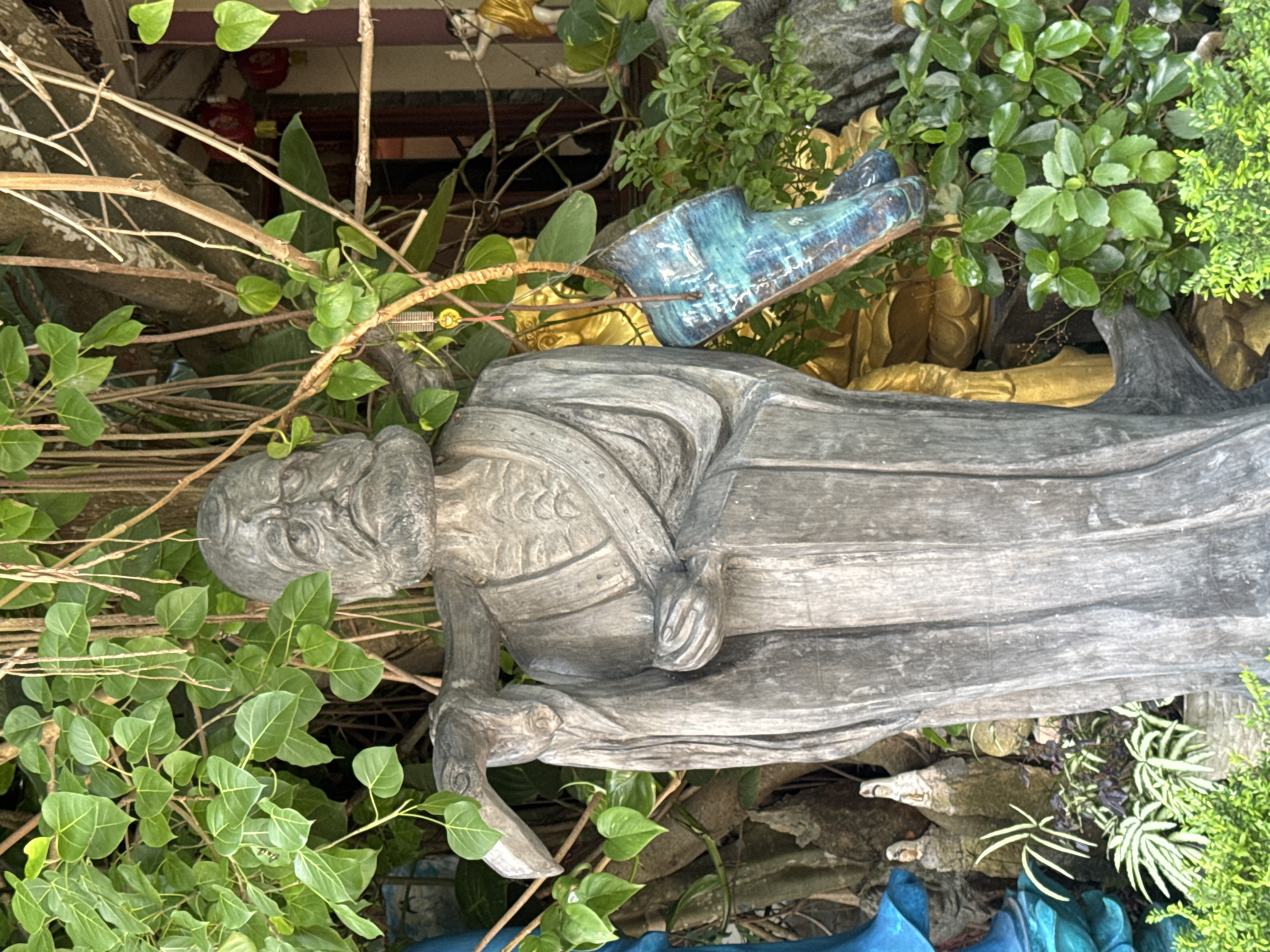 The image shows a weathered, grey stone statue of an elderly man with a long beard, wearing robes, and carrying a staff over his shoulder. The statue is surrounded by lush green foliage and trees. In the background, a golden statue is partially visible, along with two small, white statues placed on rocky outcrops. A vibrant blue ceramic piece hangs from a branch near the main statue. The overall setting suggests a garden or temple complex. This scene is located at the Quan Am Pagoda in Da Lat, Vietnam.