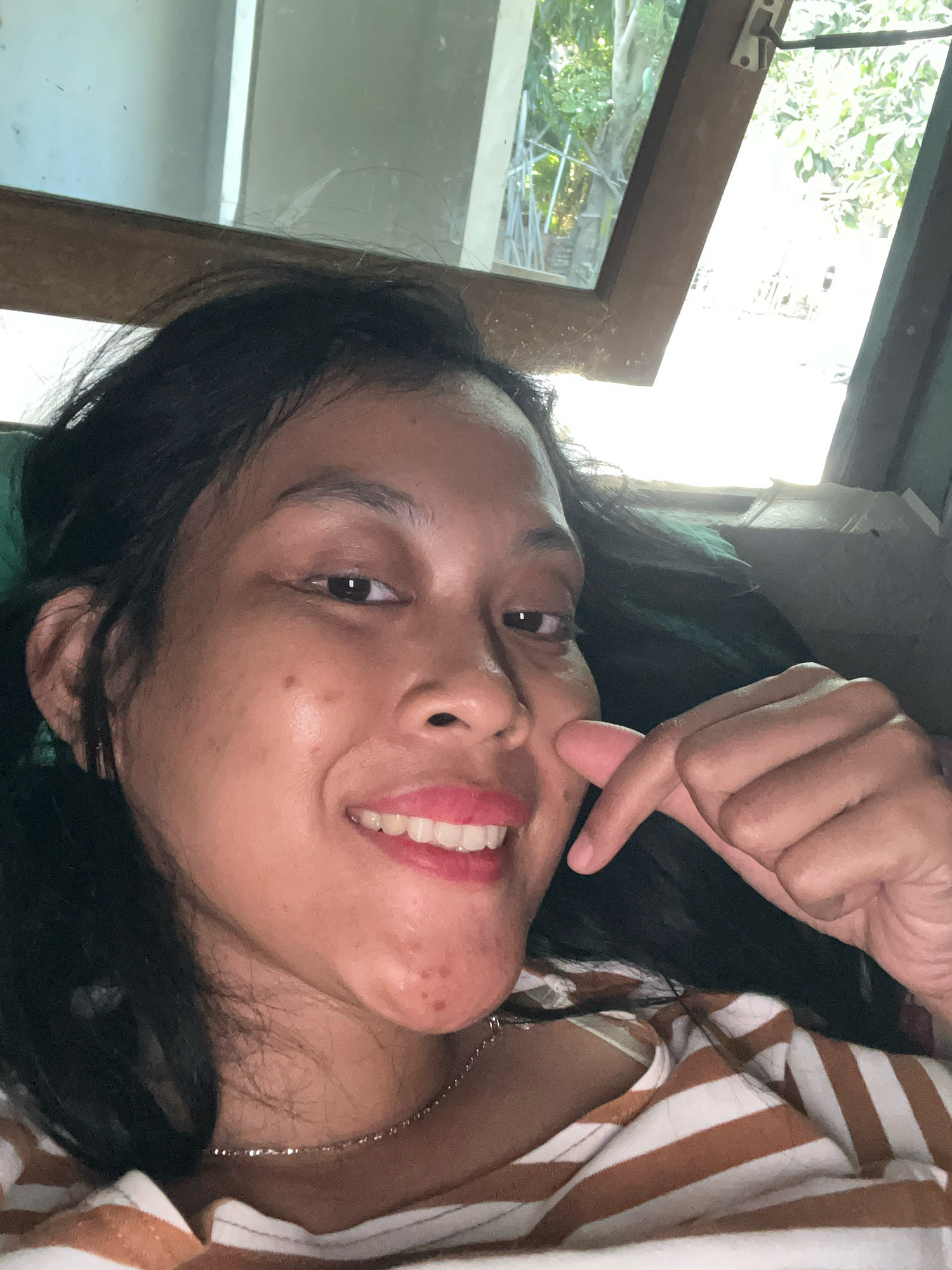 In Kecamatan Pati, Indonesia, a young woman with dark hair and light skin is taking a selfie. She is lying down, with her head tilted and her face angled towards the camera. She is smiling, with her lips parted to reveal her teeth. Her eyes are open, and she is looking directly at the camera. Her right hand is raised, and her index finger is pointing towards her cheek. She is wearing a striped shirt. In the background, there is a window with a view of trees and a building.