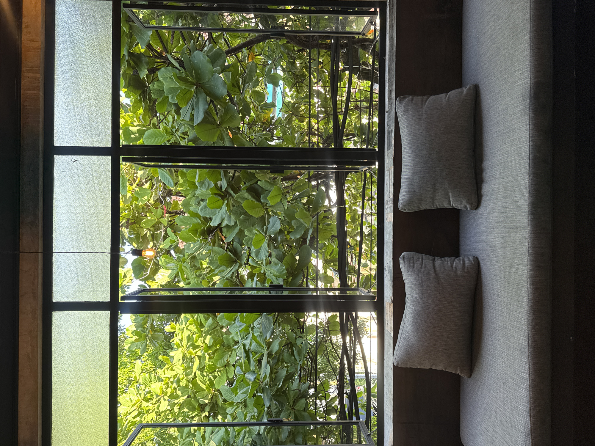 The image depicts an interior scene, looking out a large window framed by dark metal. The window is divided into multiple panes, with the lower section of three panes open, revealing lush green foliage of trees outside. Sunlight filters through the leaves, creating a dappled effect. Above the open panes, the upper portion of the window consists of frosted glass, diffusing the light. A single, exposed light bulb hangs from a cord near the top of the window. In the foreground, a dark wooden ledge runs along the base of the window. On this ledge rests a grey upholstered sofa with two matching grey throw pillows. The overall impression is one of a tranquil seating area with a view of nature. The specific location is identified as Kuta, Indonesia.